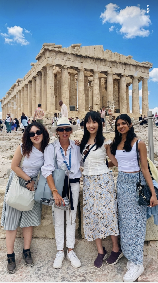 Four women standing together in front of the Parthenon in Athens, Greece, with other tourists in the background on a cloudy day.