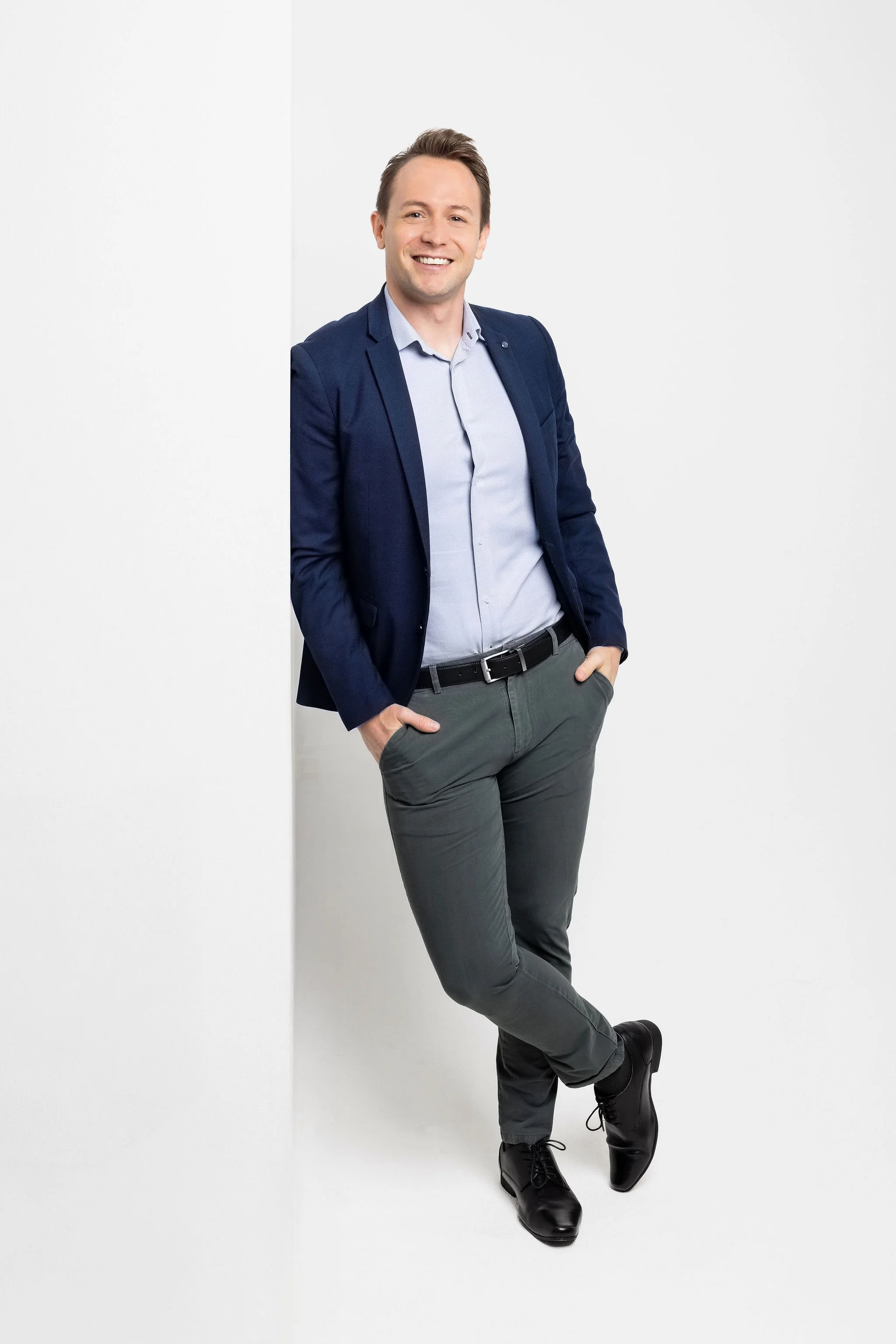 A young man in a business casual outfit smiling and leaning against a white wall.
