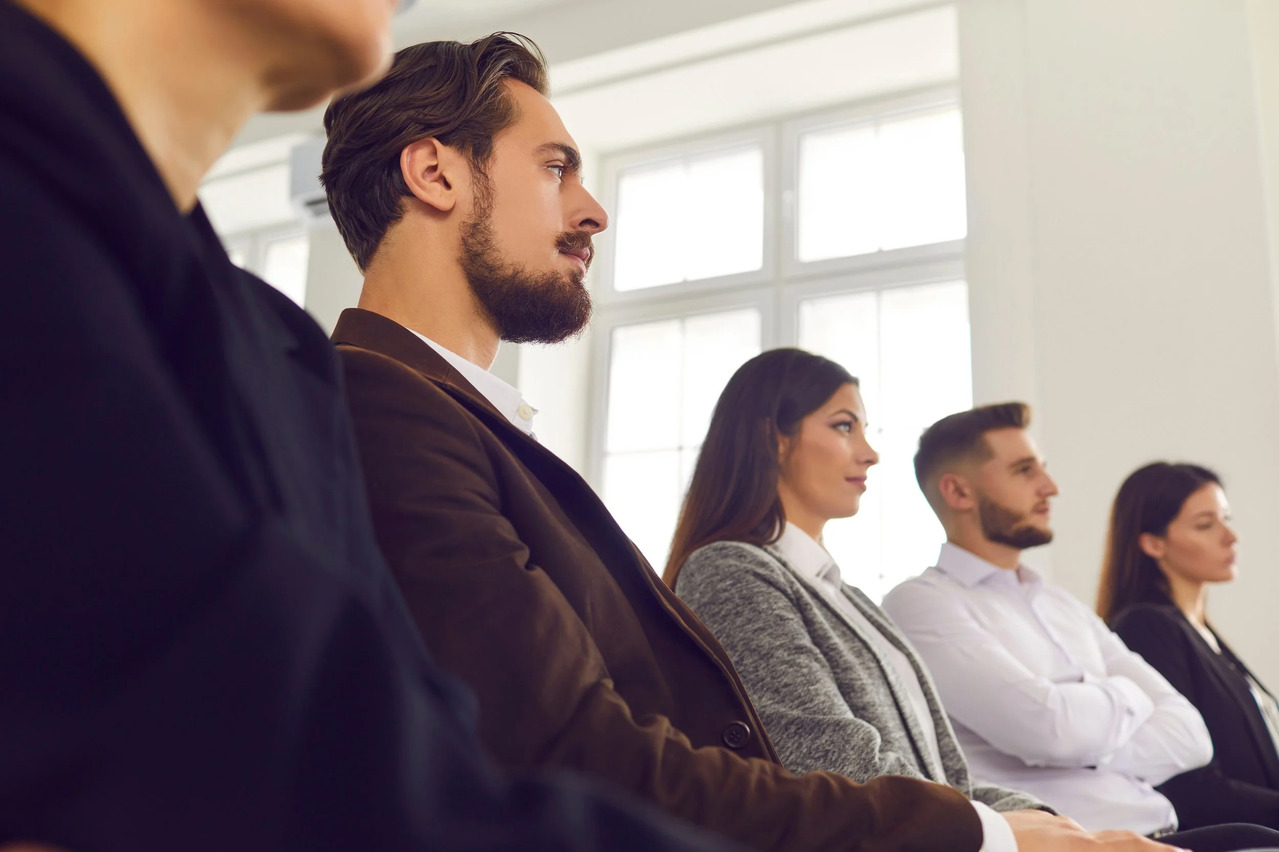 A group of five young professionals sitting in a row, attentively listening during a meeting or conference in a bright, modern office space.