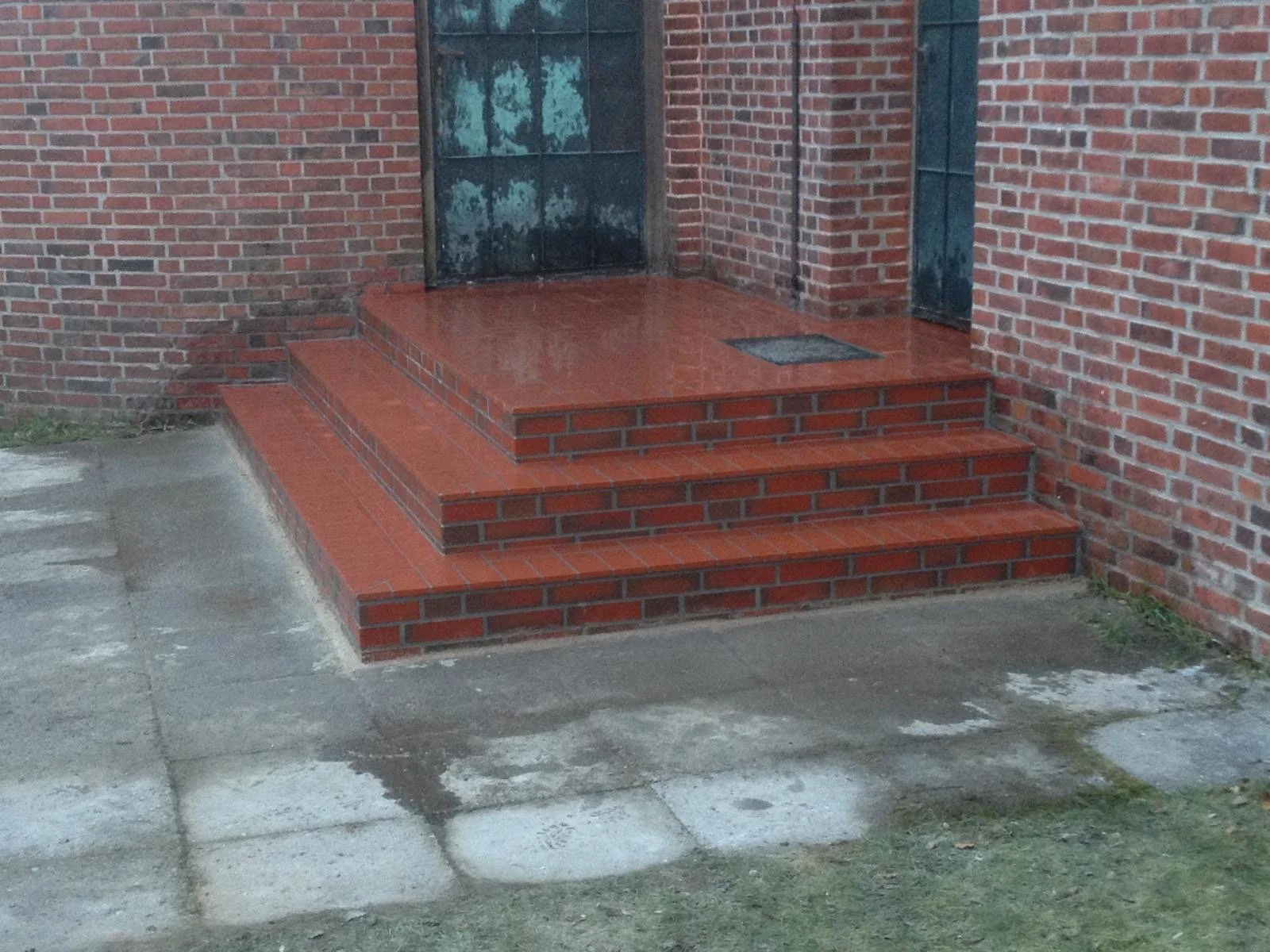 Rote Ziegeltreppen vor einer roten Backsteinwand und einer schwarzen Tür, auf nassem Betonboden.