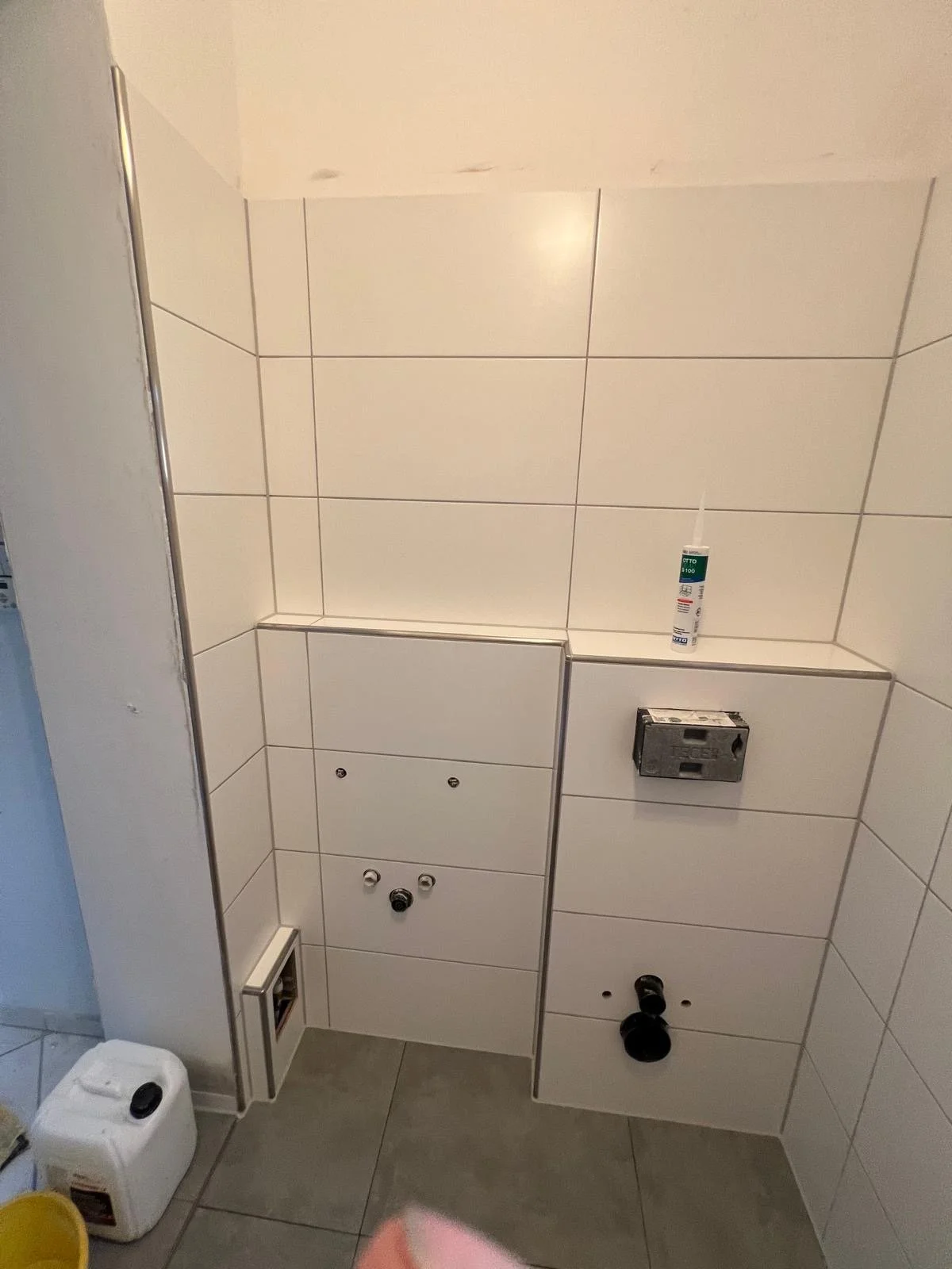 Badezimmerwand mit weißen Fliesen, Waschmaschinenanschluss, Dichtstoff und Rohrleitungen ohne Toilette oder Waschbecken.