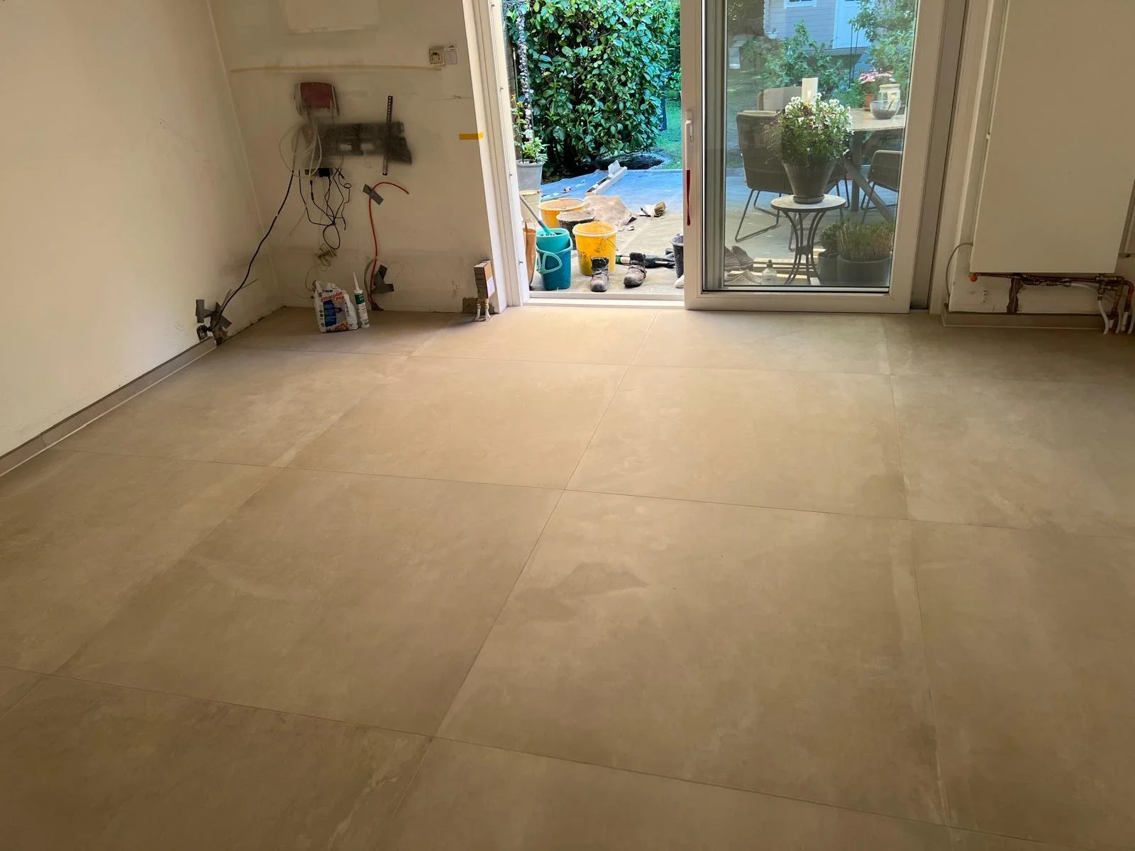 Innenraum eines Hauses während der Renovierung mit offenem Ausgang zum Garten, unvollständige Küche, Böden werden verlegt, im Garten sind Gartenwerkzeuge und Pflanzen sichtbar.