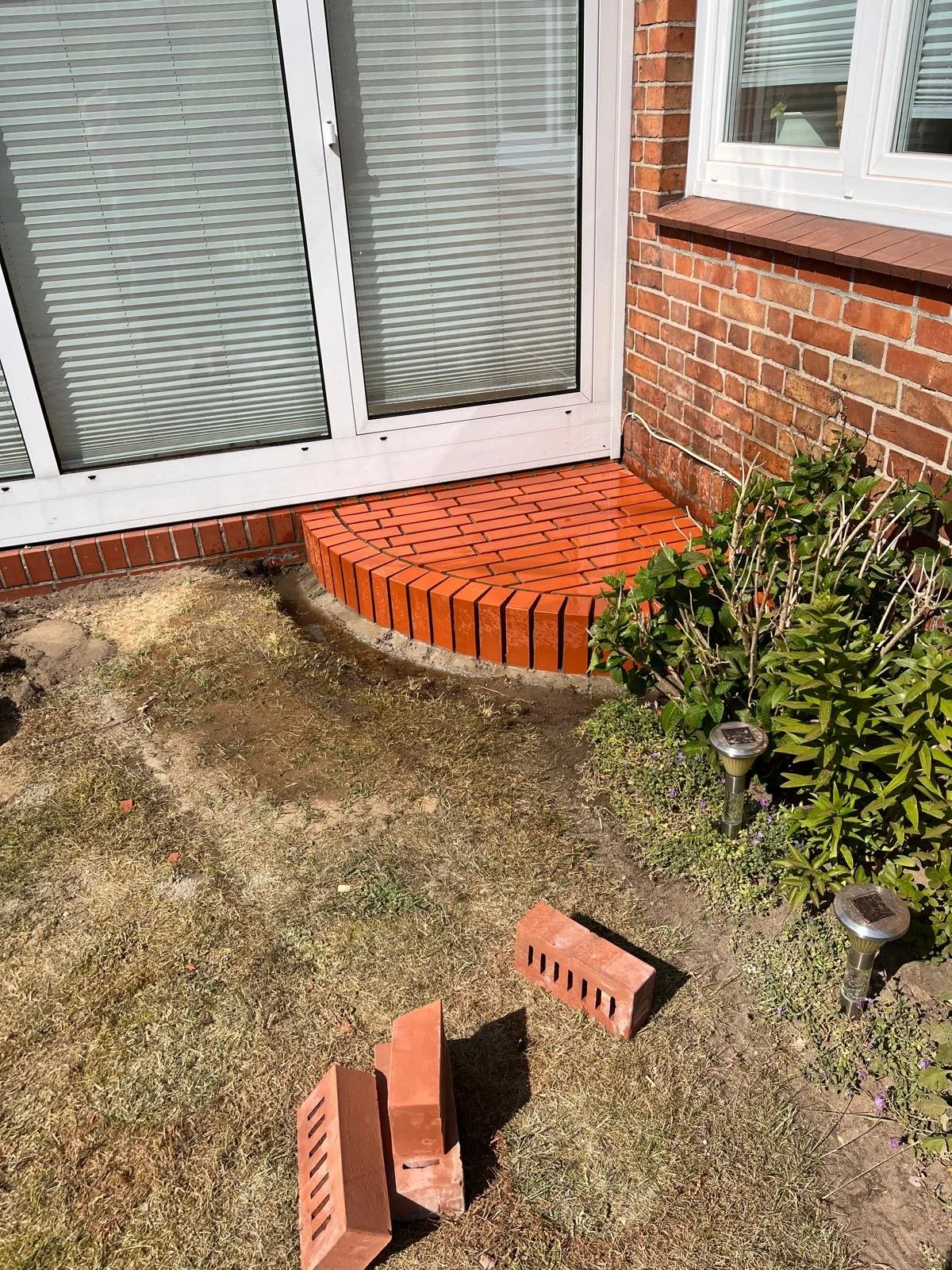 Ein frisch verlegter roter Ziegelsteg vor einer Terrassentür, umgeben von Gartenpflanzen und elektrischen Gartenleuchten auf einem Kies- und Erdboden.