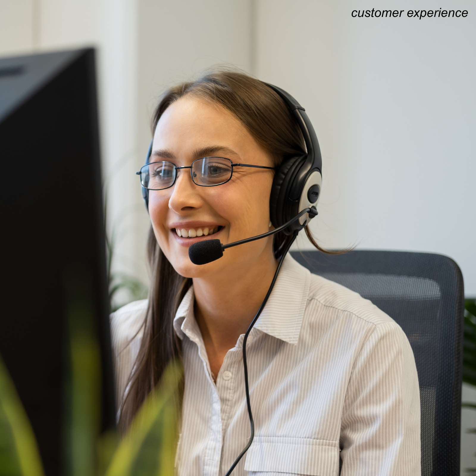 A woman wearing glasses and a striped white shirt, smiling and looking at a computer screen. She has a headset with a microphone on, indicating she is likely working in customer service or a call center. There is a plant in the foreground and a plain wall in the background.