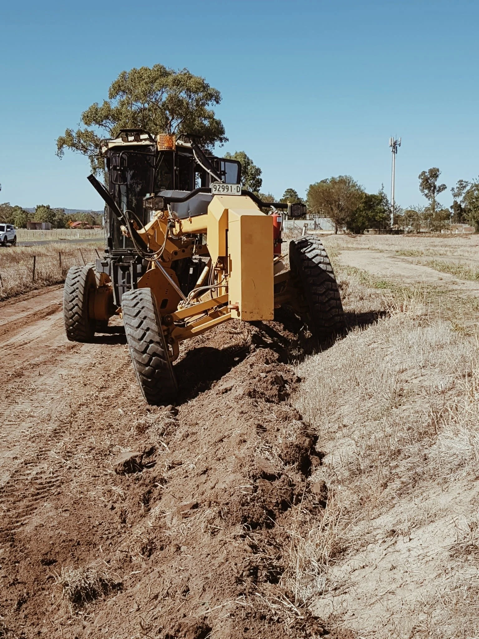 A yellow construction grader on a dirt field with trees and a water tower in the background under a clear blue sky.
