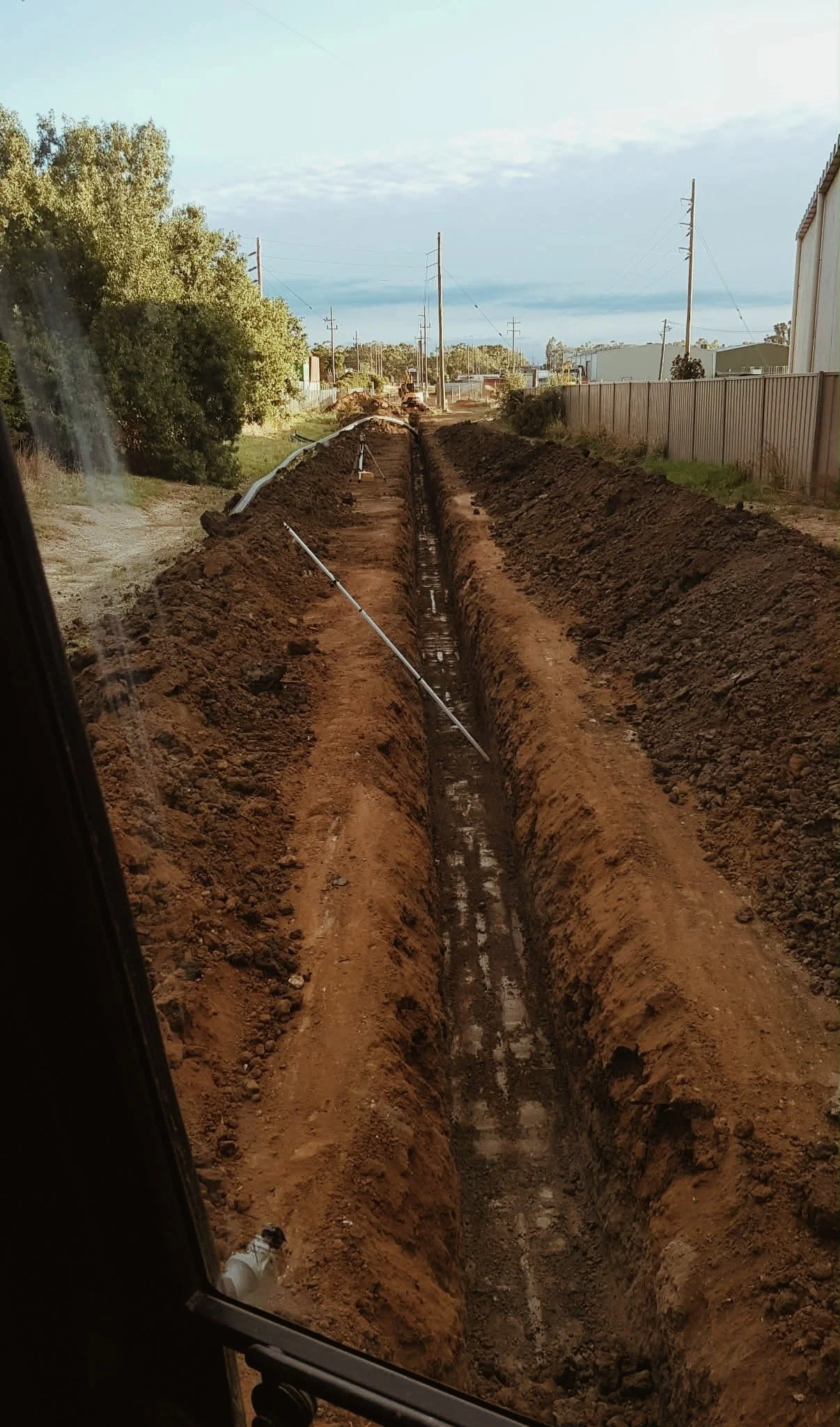Construction site with a deep trench for installing underground pipes or cables, with dirt piles on either side and utility poles along the street in the background.