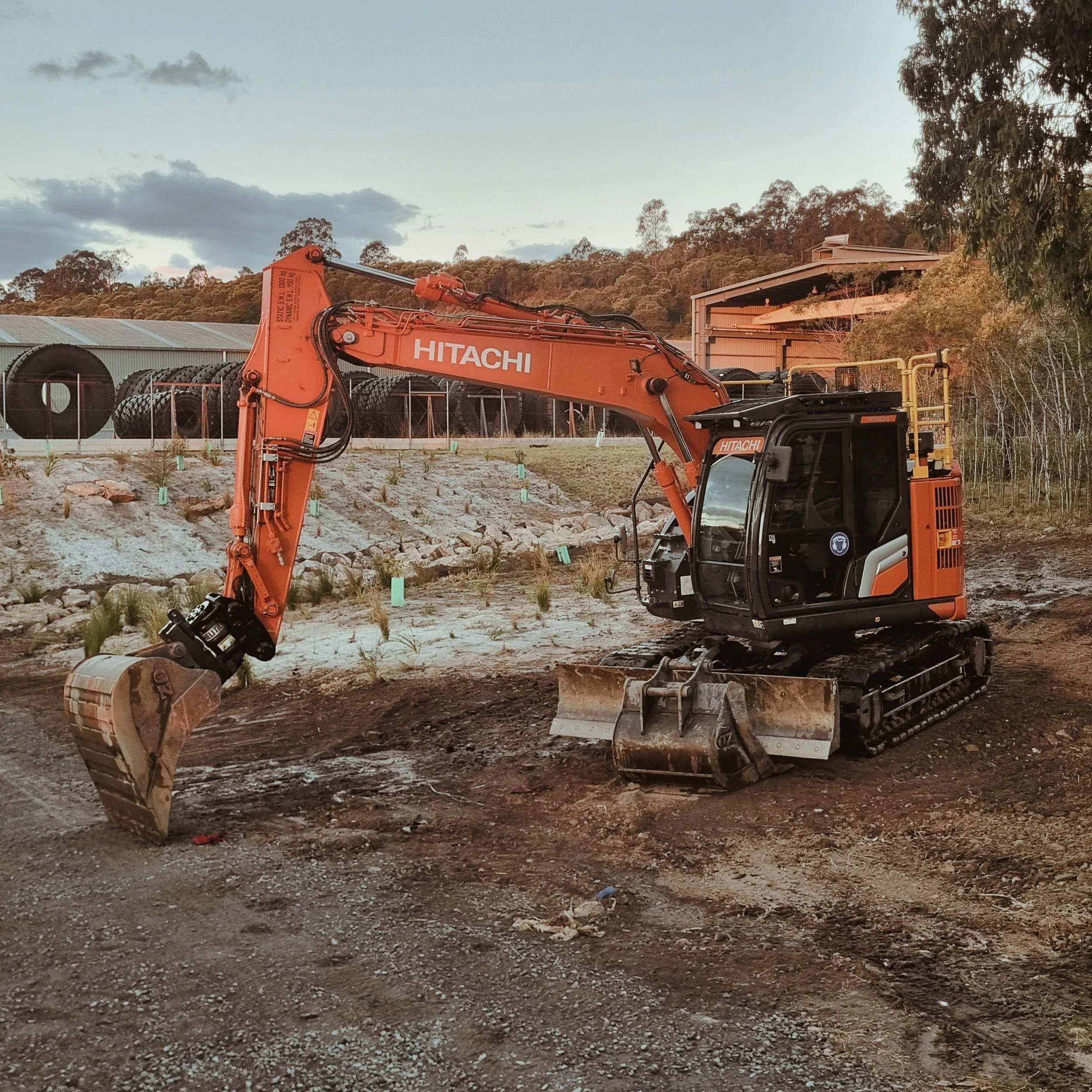 Construction site with an orange Hitachi excavator, large pipe rolls, and a partially built structure in a rural area with trees and a cloudy sky.