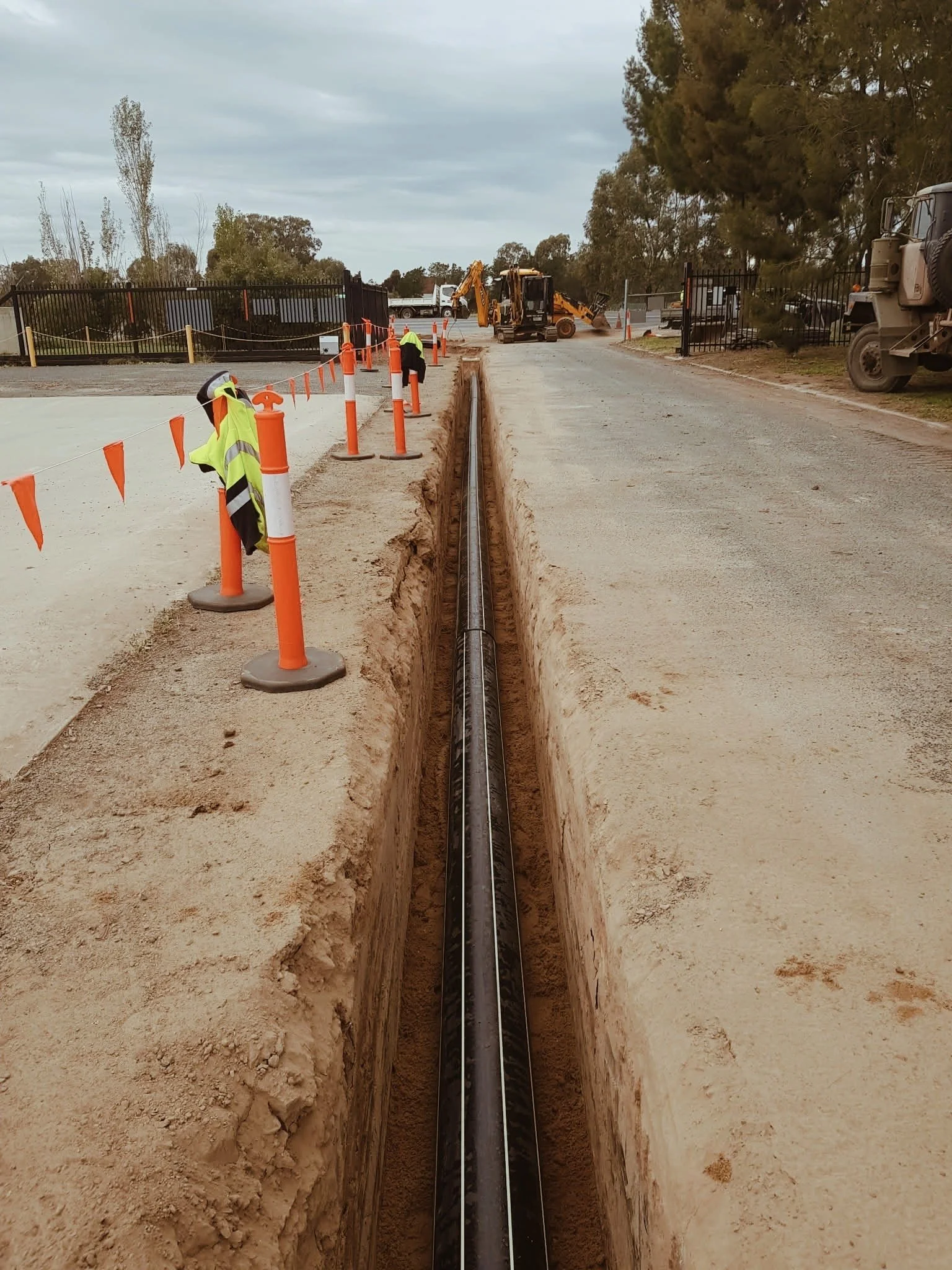 Construction site on a road with a large trench, a pipe being installed, orange safety cones, and machinery in the background.