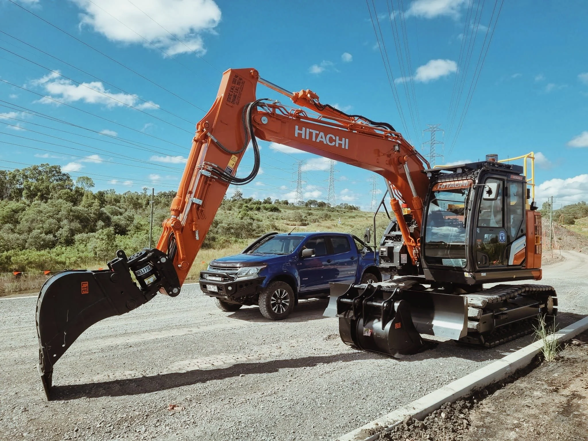 An orange Hitachi excavator on a construction site next to a blue pickup truck, with multiple power lines and a green hilly landscape in the background, under a partly cloudy sky.