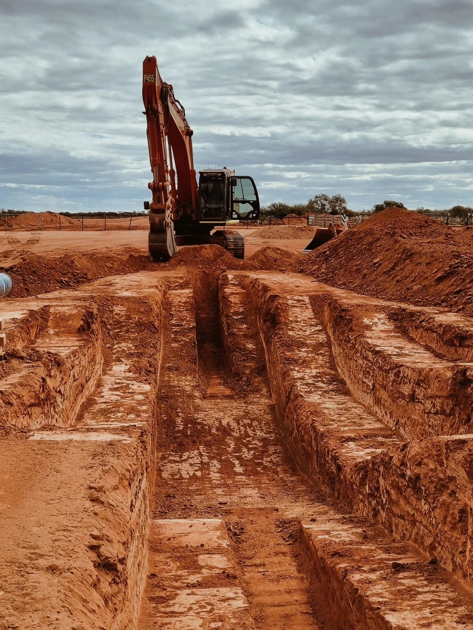 Construction site with a small excavator digging trenches in reddish-brown dirt under a cloudy sky.