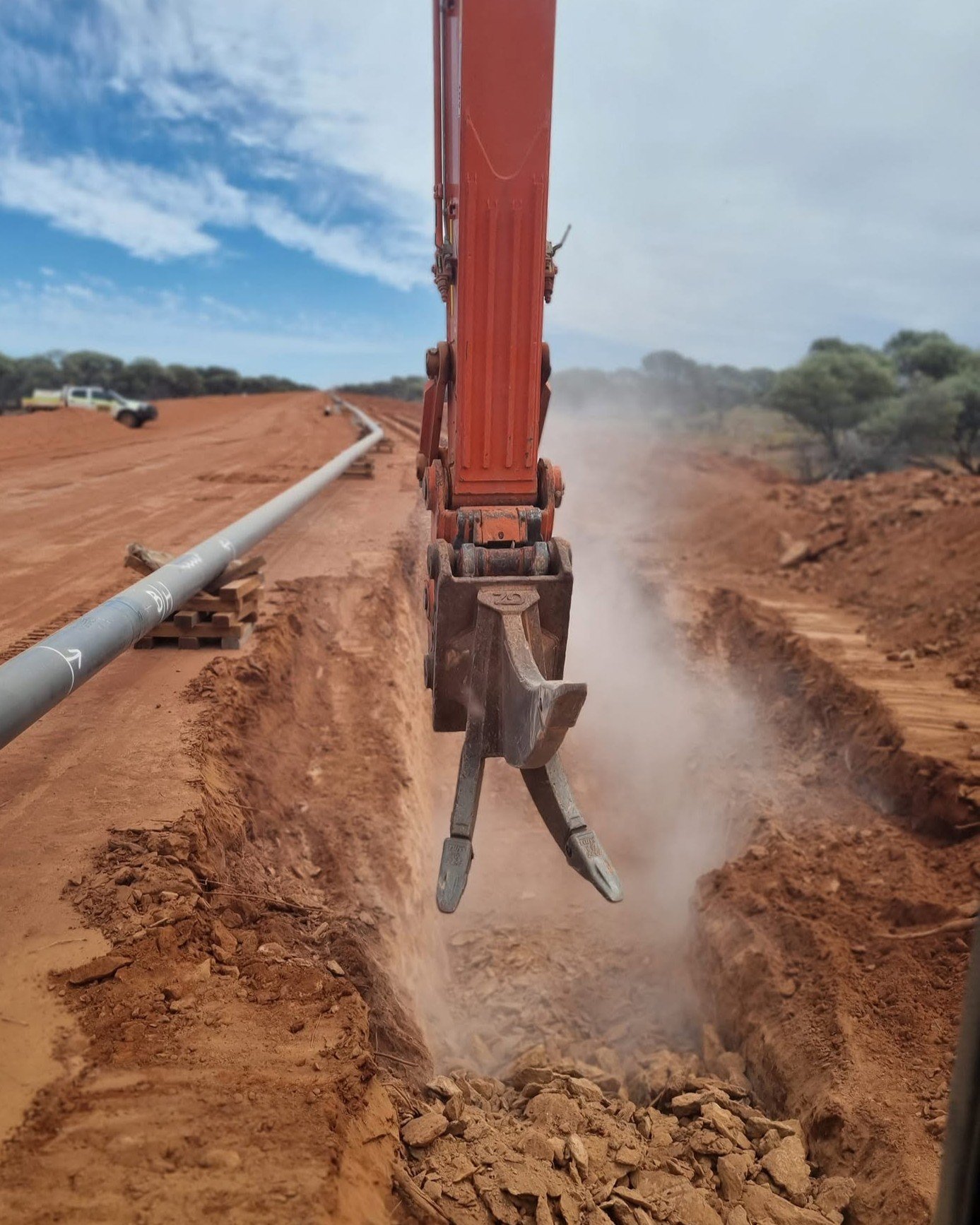 Close-up of a construction excavator digging a trench on a dirt road during daytime, with a pipe running along the trench, and trees and sky in the background.