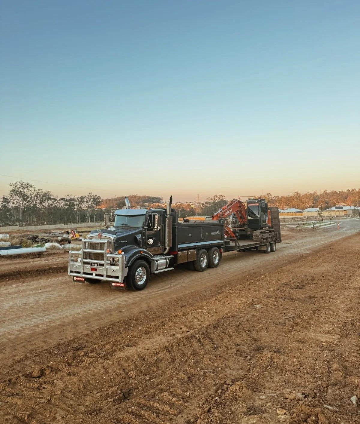 A large black truck with a bull bar is pulling a flatbed trailer carrying an orange excavator on a dirt road at sunset. In the background, there are construction materials, pipes, and structures.