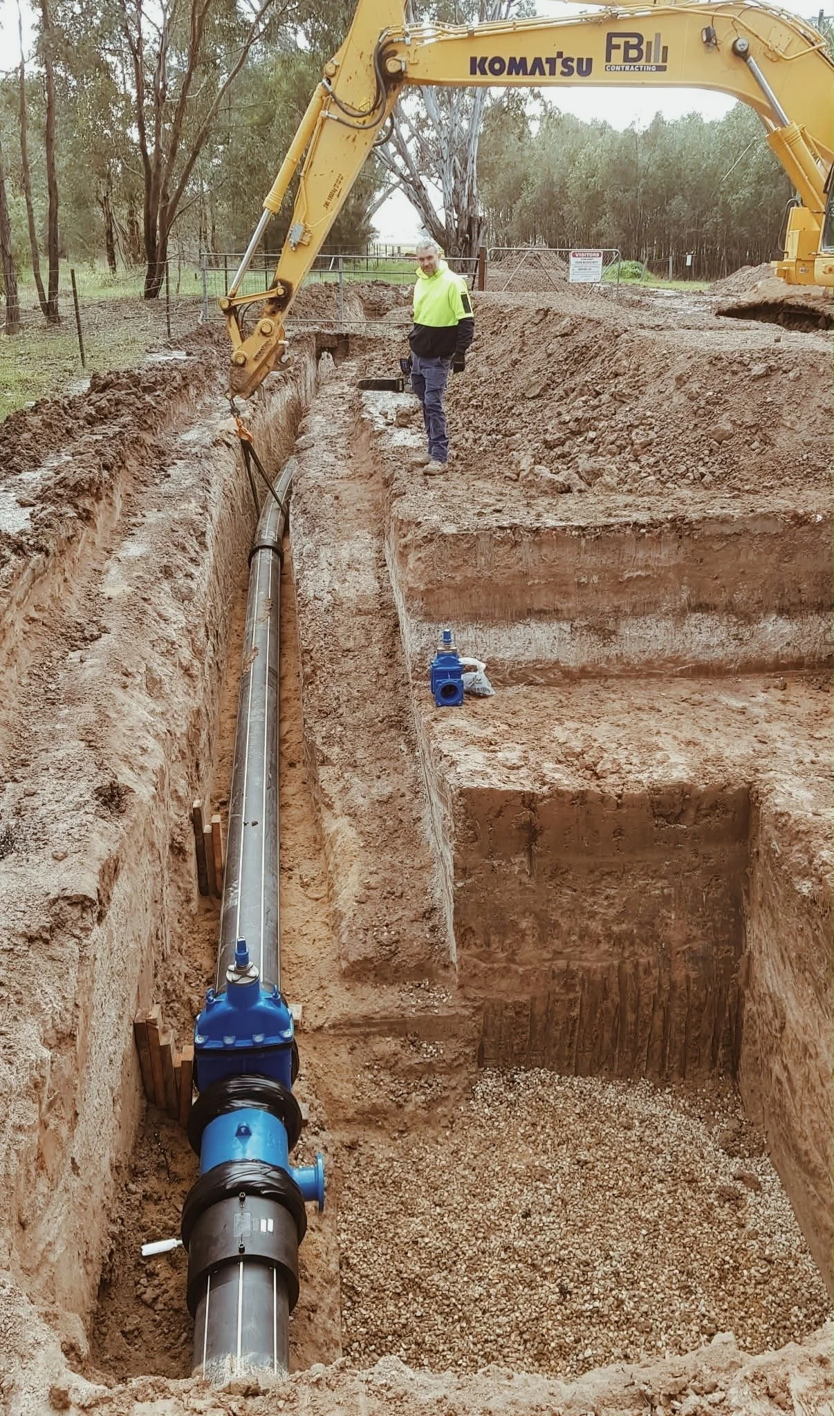A construction worker stands in a long, deep trench with a large Komatsu excavator. The trench contains a pipeline with blue fittings, and danger signs are visible in the background. The area is surrounded by trees and open land.