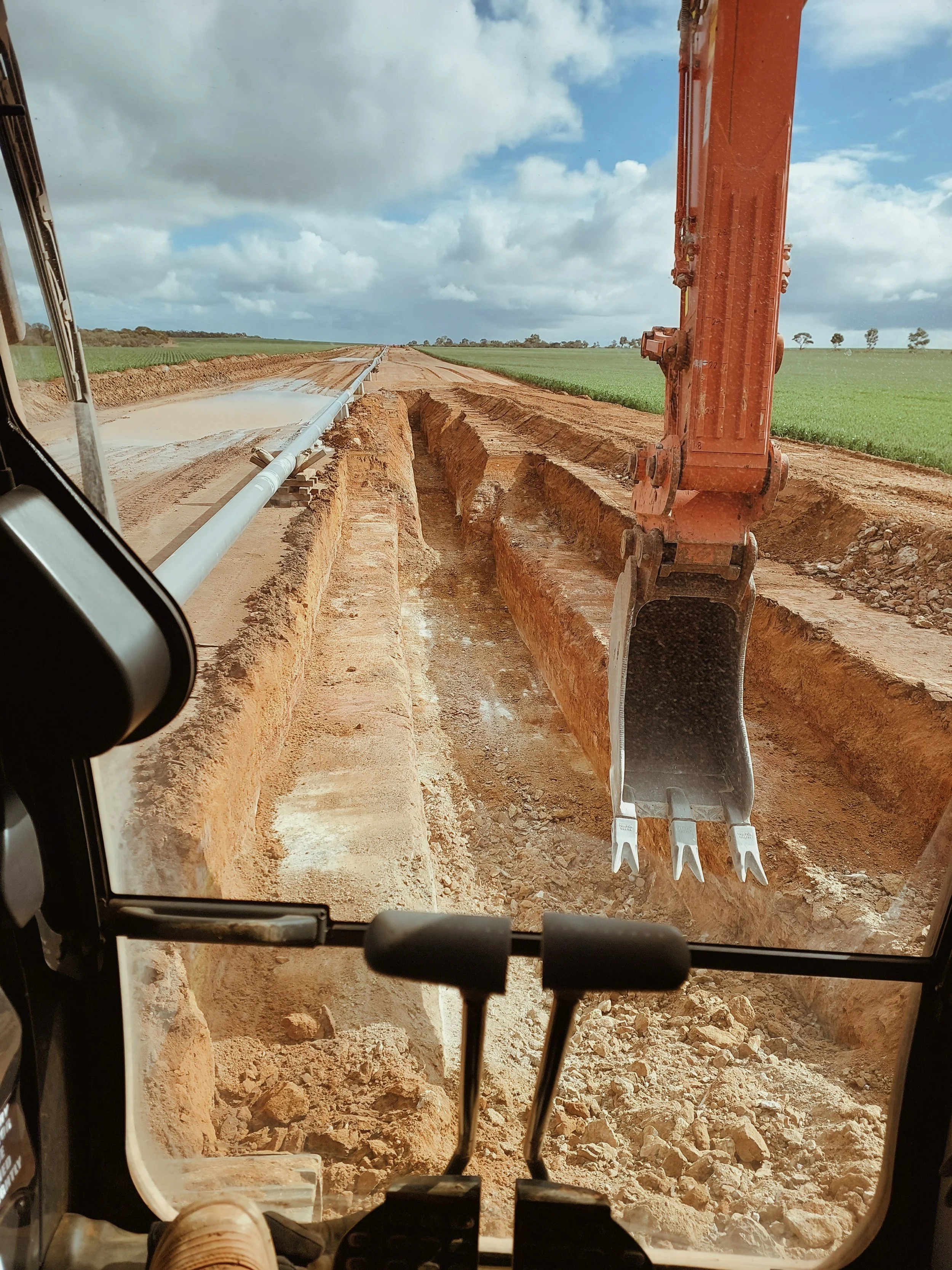 A view from inside a construction or excavation vehicle cabin showing a large excavator arm and bucket digging a trench for pipeline installation in a rural area with green fields and cloudy sky in the background.