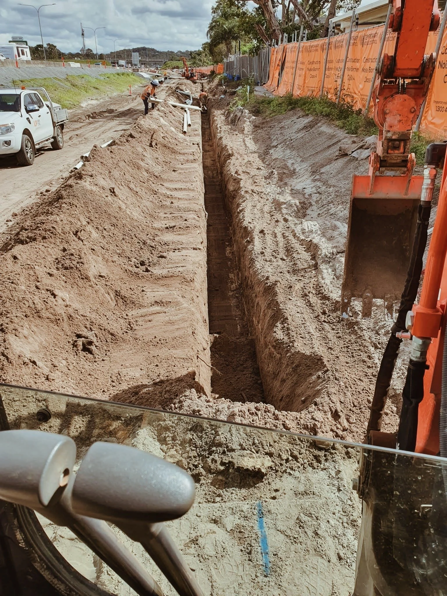 Construction workers are working in a trench on a sandy roadside, with construction equipment and orange safety barriers on the side.