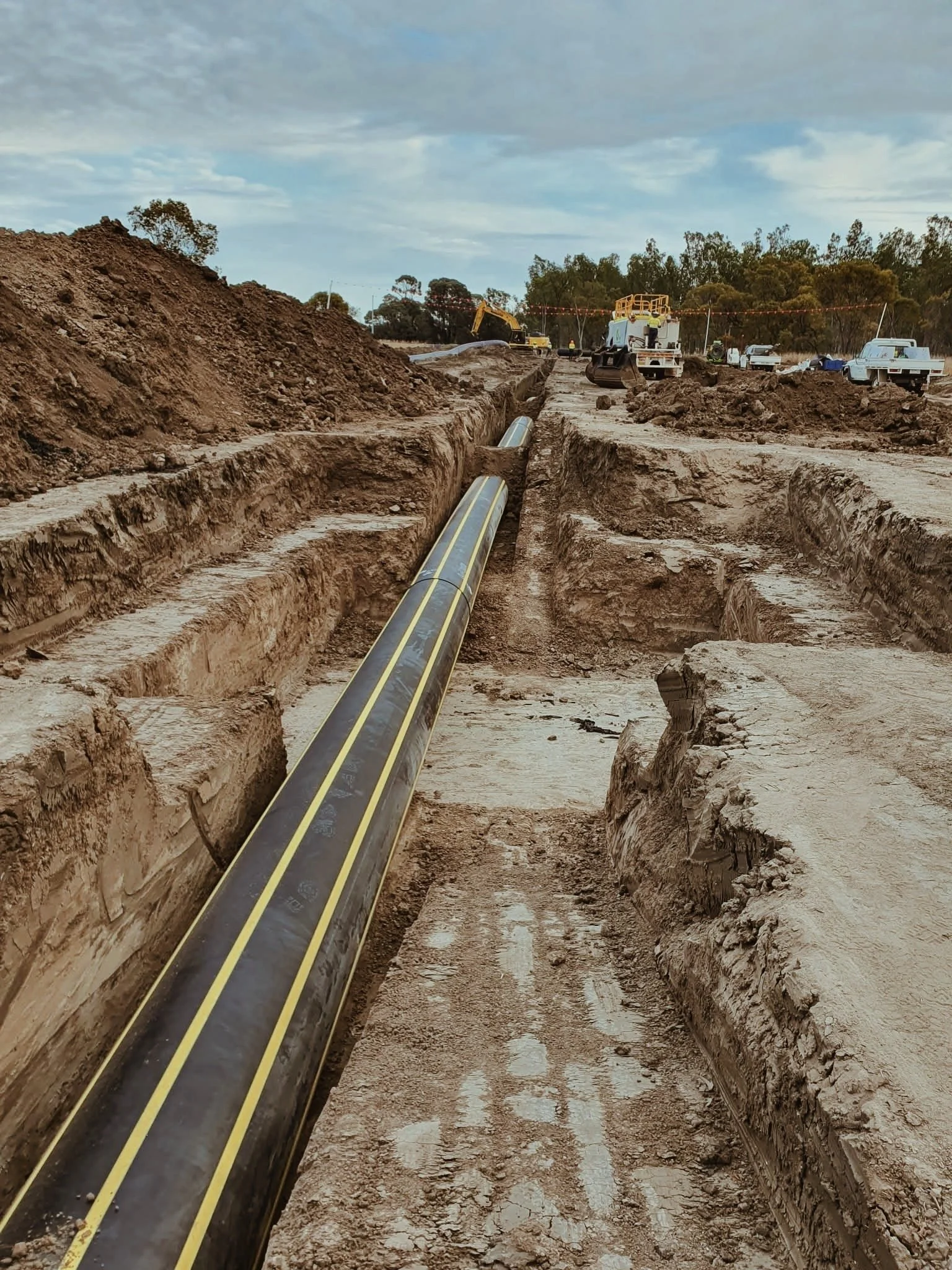 Construction site with a pipeline being installed in a trench, heavy machinery and workers in the background.