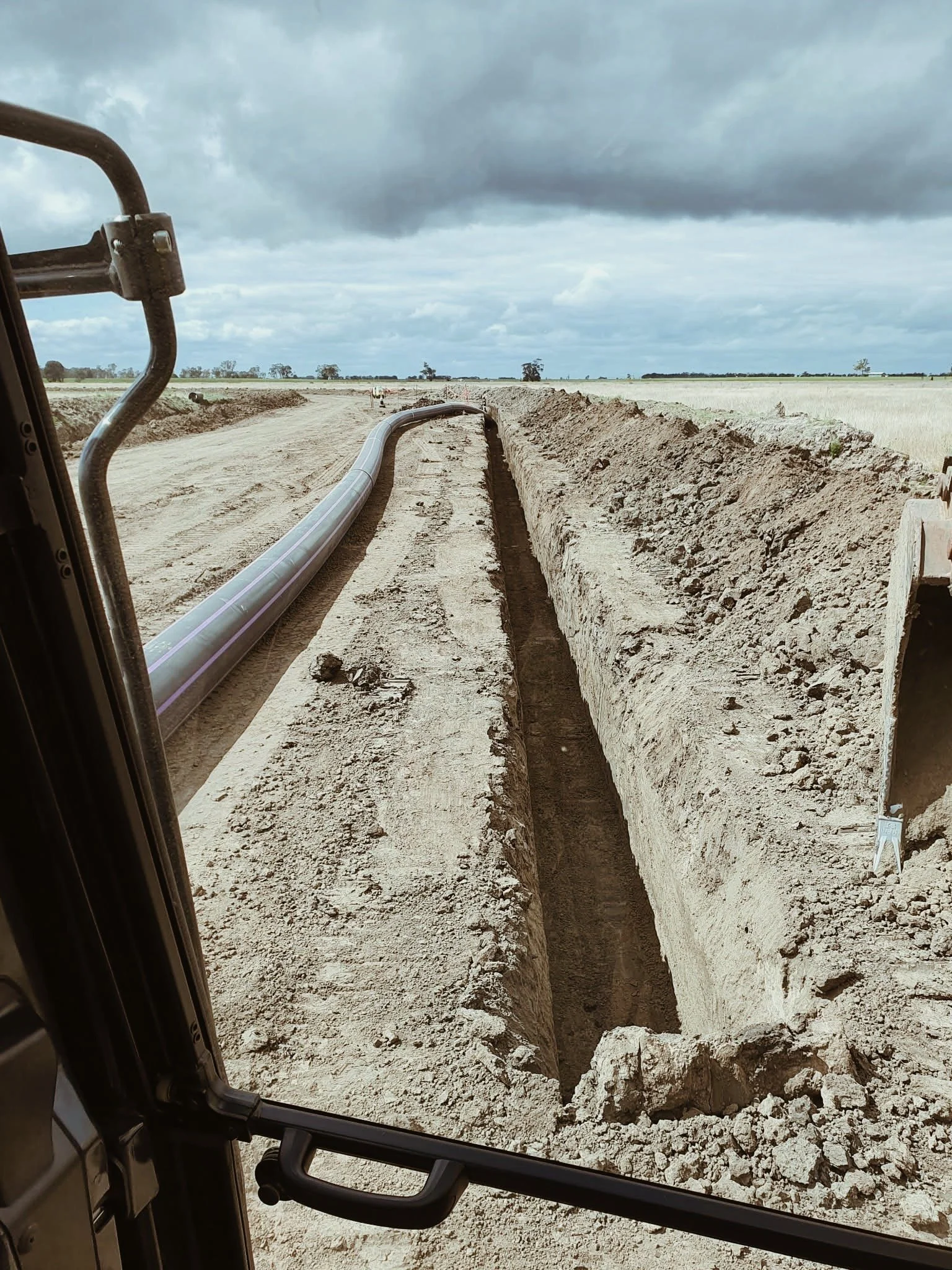 View from inside construction equipment of a dirt road with a trench dug along its side and large pipes laid in the trench. Overcast sky with dark gray clouds above a rural landscape.