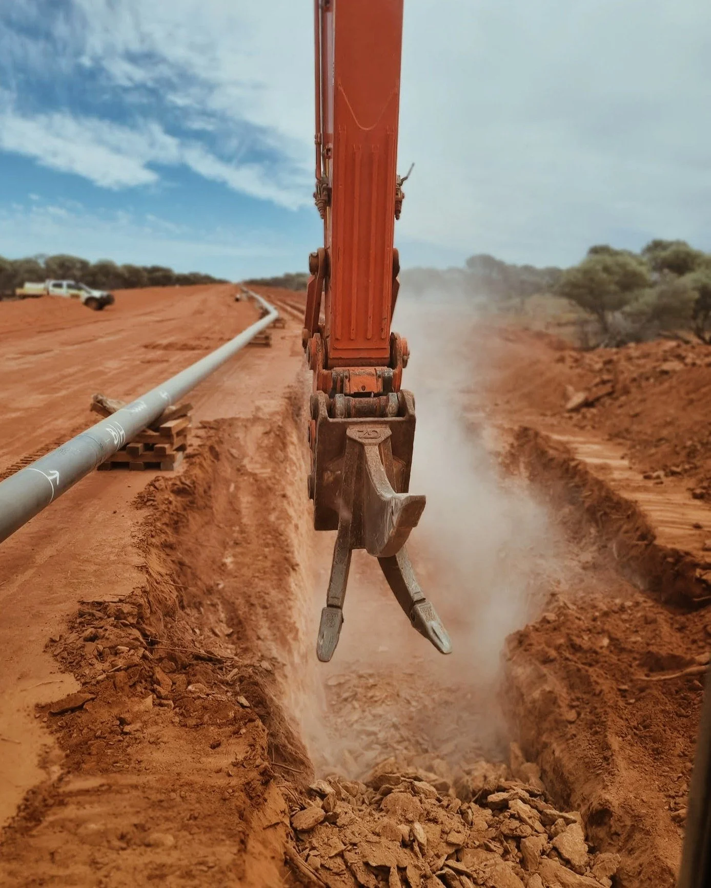 View of an orange excavator arm digging a trench in dry, reddish soil, with a pipeline running through the trench and dust rising from the operation. There are trees and vehicles in the background under a partly cloudy sky.