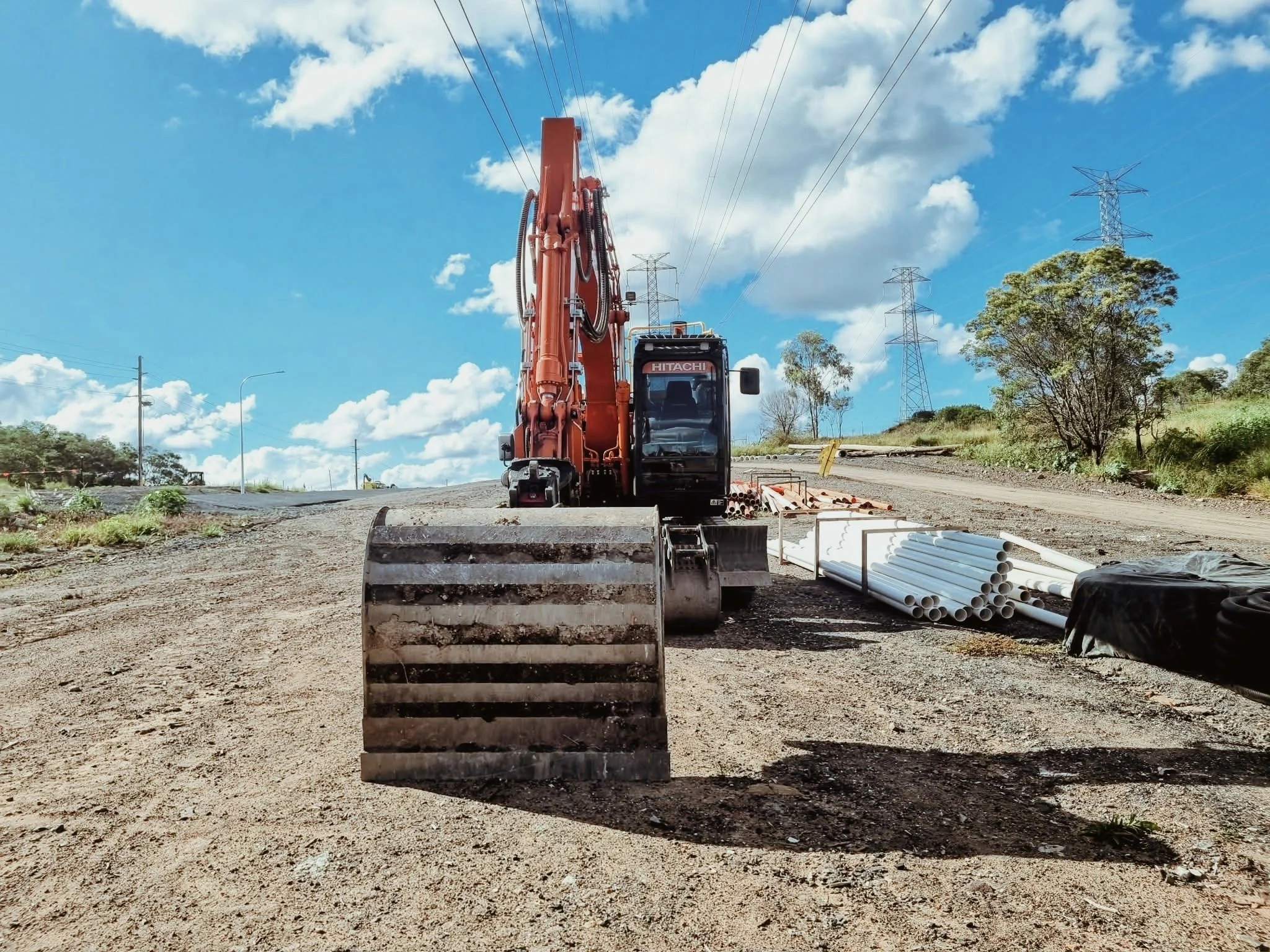 Construction site with an orange excavator, pipes, and soil, under a blue sky with clouds and power lines.