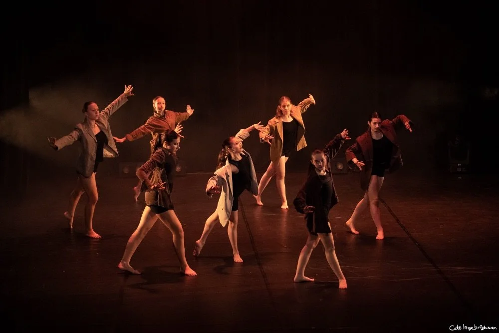 Ti unge jenter som danser på en scene, kledd i forskjellige jakker og svart treningstøy, under en teaterforestilling.