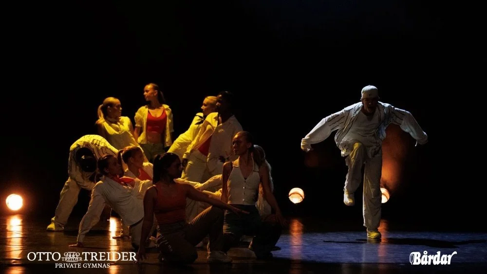 Scenekunst med flere dansere på scenen, noen som sitter, noen som står, og én som hopper i midten, opplyst av scenebelysning.