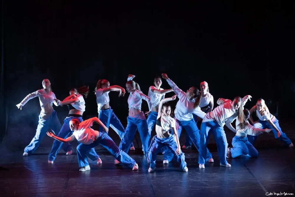En gruppe unge kvinner som danser på en scene i uniformer, kledd i hvite topper og blå bukser, under en forestilling.