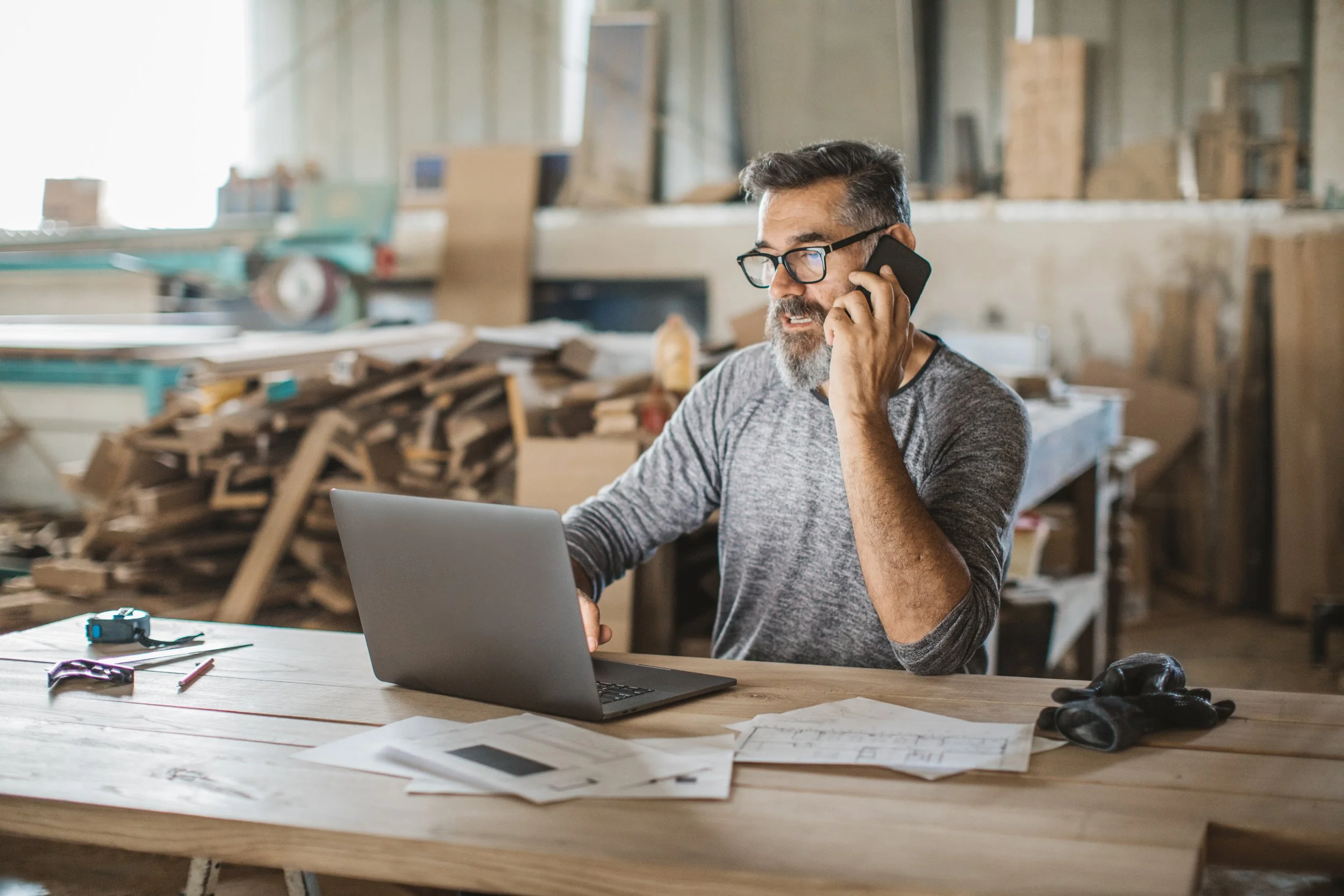 A man with glasses and a beard talking on a cell phone, working on a laptop in a woodworking workshop filled with wood scraps and tools.