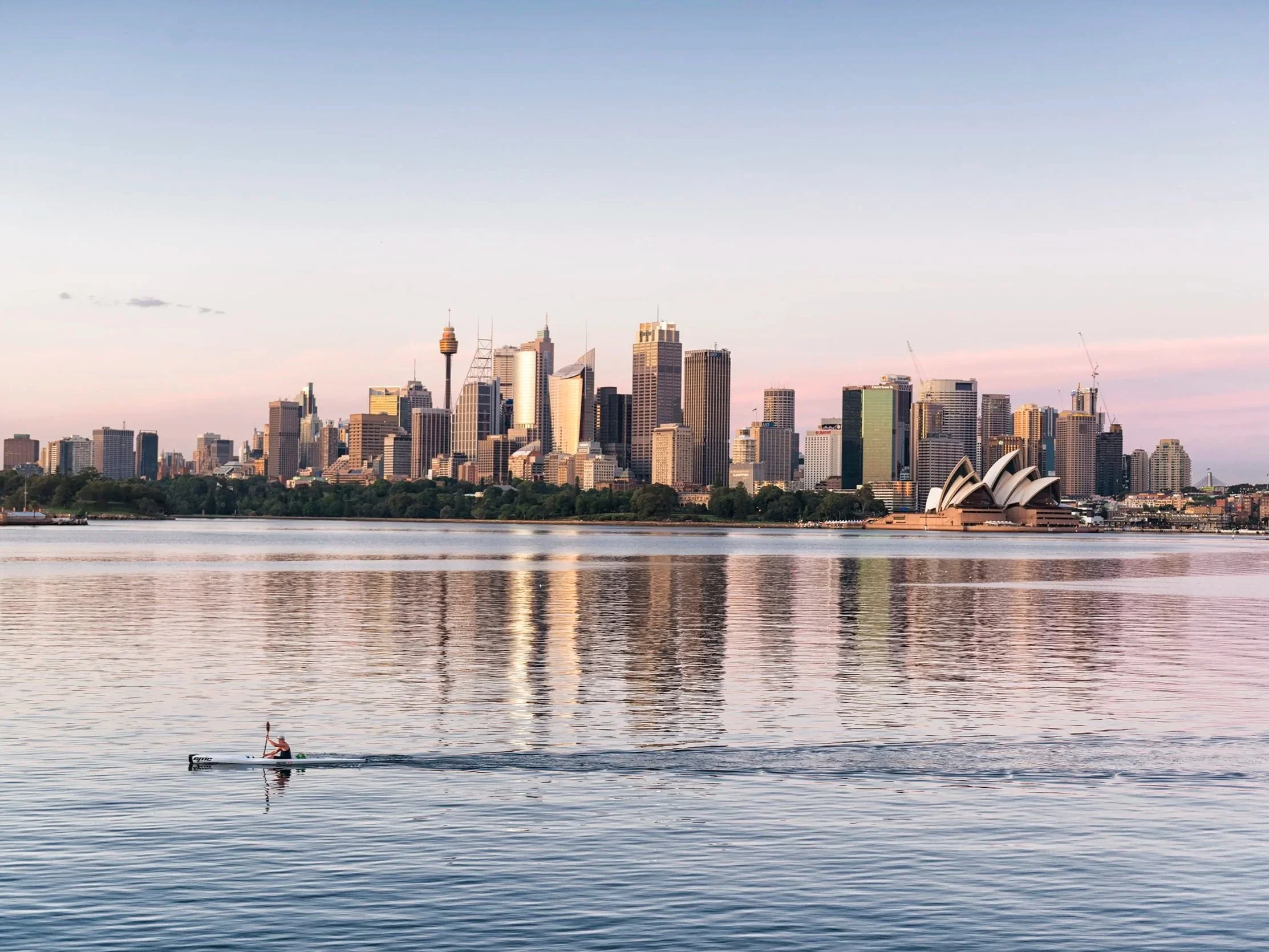 Sydney skyline with the Sydney Opera House in the foreground and a person paddling a kayak on the water.
