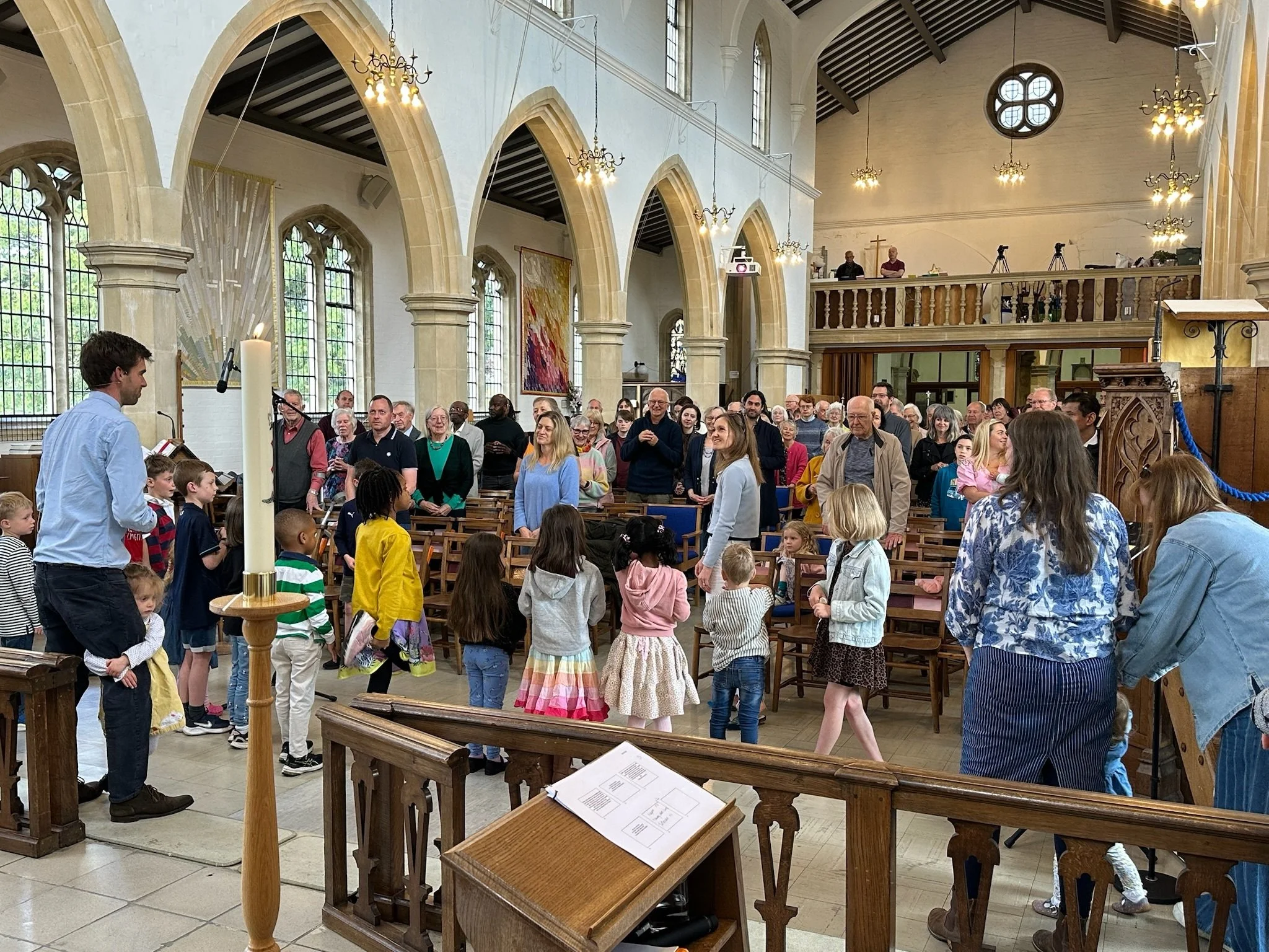 A congregation gathered inside a church with high arched windows and stone columns. Children and adults stand in the center, some singing or participating in a service. The church features wooden pews, hanging chandeliers, and an elevated balcony area in the background.