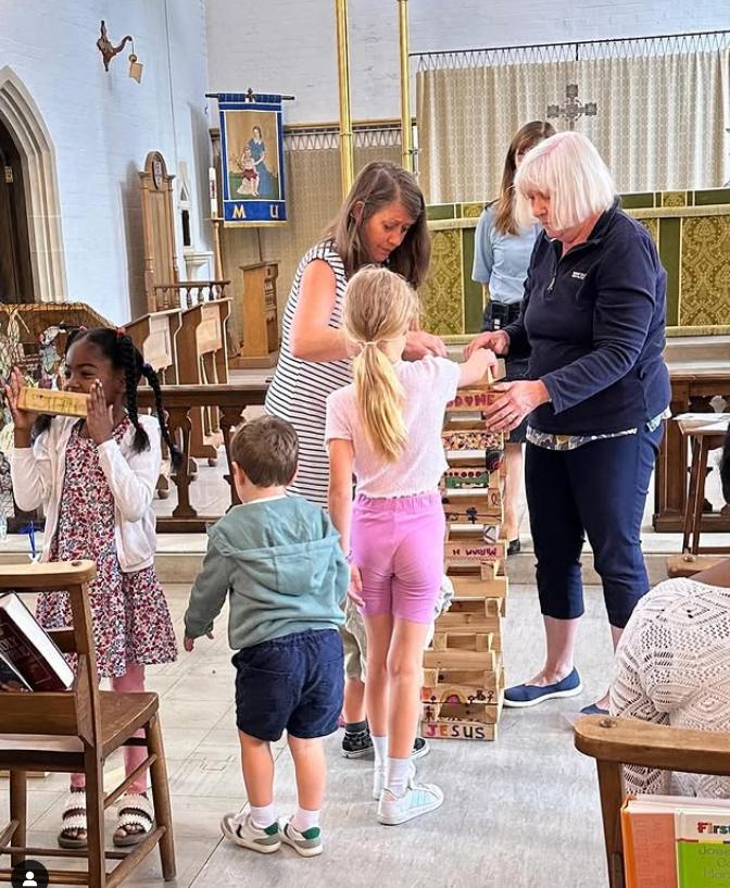 Children playing a game of Jenga inside a church, with a woman overseeing the game and other children observing, in a church interior with religious icons and banners.