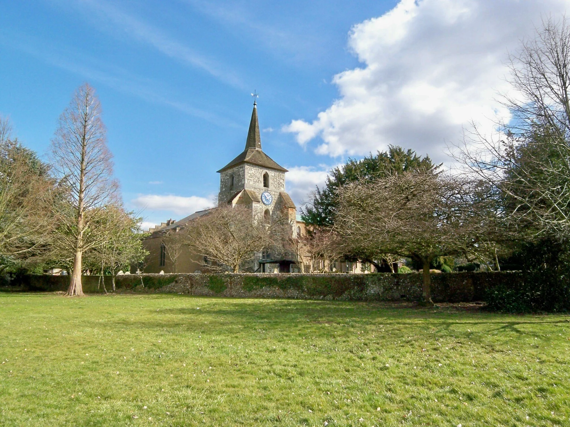 A church with a tall steeple and clock in a grassy field with trees and a blue sky with clouds.