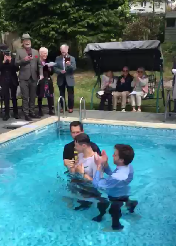 Three men are standing in a swimming pool, with one of them possibly performing a baptism or a similar ceremony. Several people are gathered on the pool deck, some taking photos with their phones and others sitting on a swing bench, watching the event.