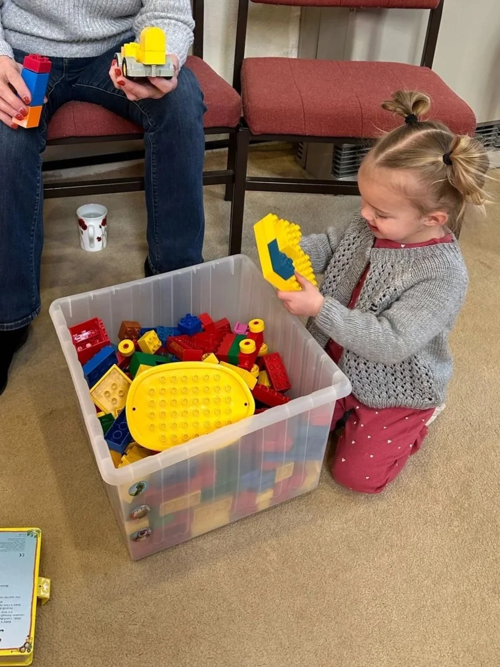 A young girl with pigtails kneeling on carpeted floor, playing with yellow, red, and blue LEGO bricks from a large plastic container, while an adult sitting nearby holds additional LEGO pieces.