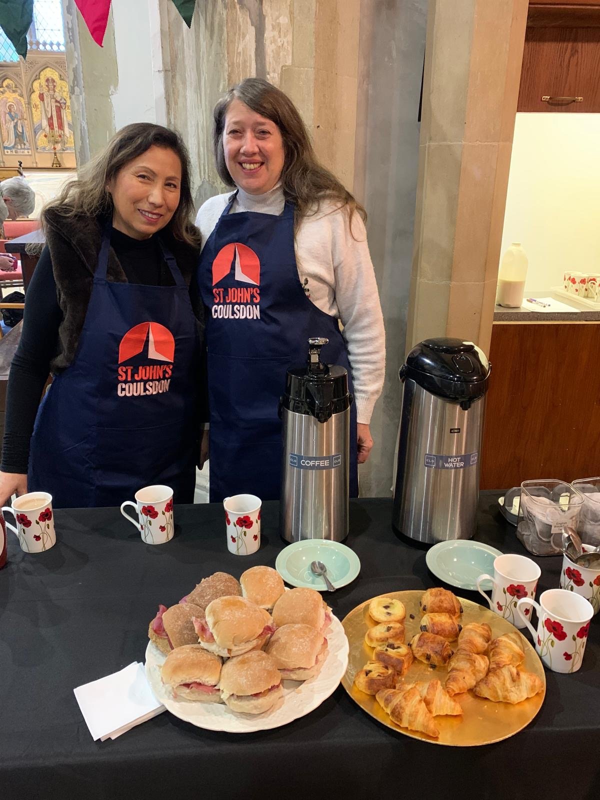 Two women smiling at a table set with breakfast items, wearing matching blue aprons with a red and white logo, in a church or community hall setting.