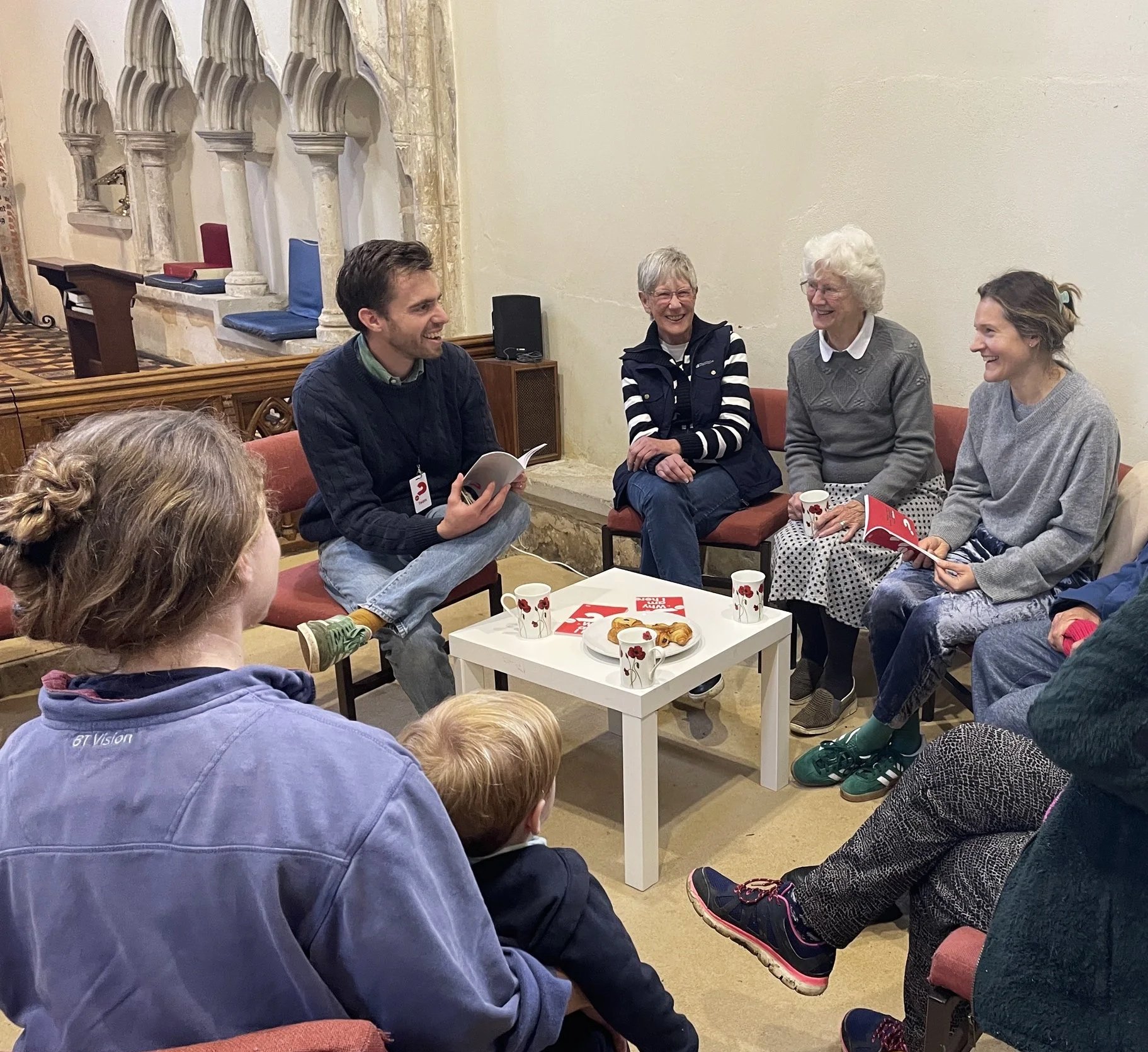 Group of eight people seated around a small table in a cozy room, engaged in a conversation, enjoying coffee and snacks, some holding booklets, with a church or historical building interior in the background.