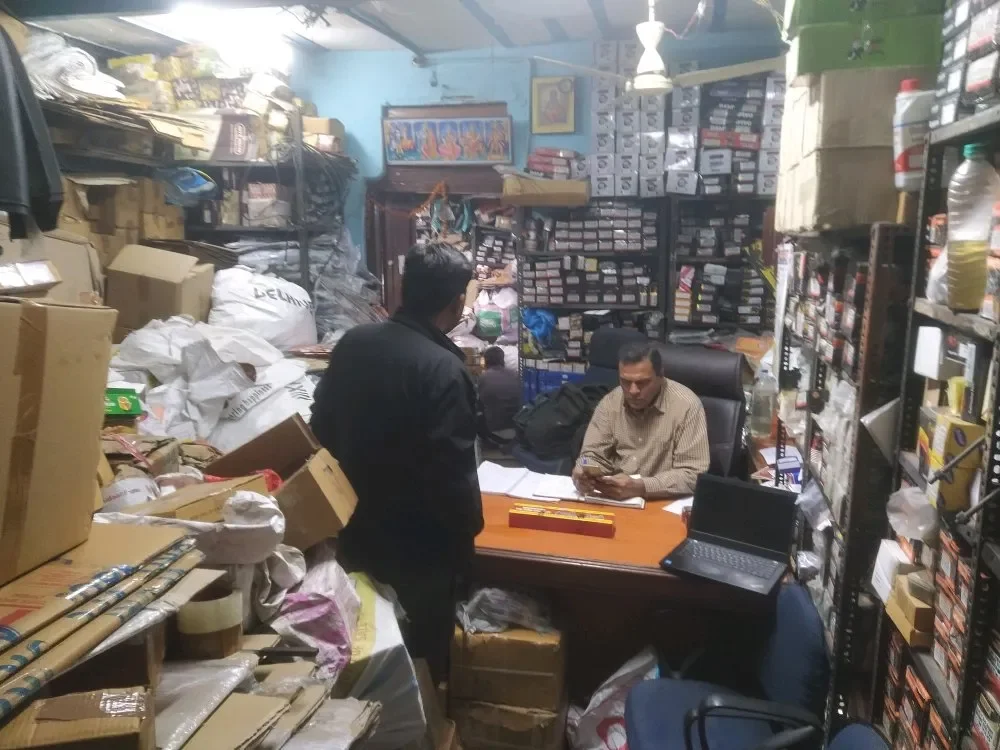 Two men in an office filled with shelves of boxes and packages, one seated at a desk using a phone, the other standing and talking to him, surrounded by piles of paper and packaging materials.
