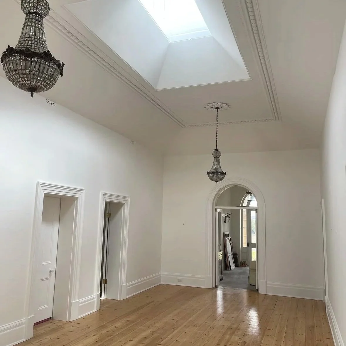 heritage house restoration and repaint. Empty room with white walls, hardwood floors, ornate crown molding, two vintage chandeliers, a skylight, and an arched doorway leading to another space with large windows.