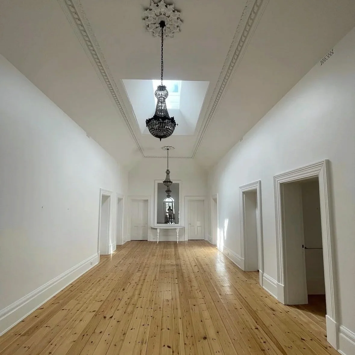 heritage house restoration and repaint. Empty, spacious hallway with white walls, wooden floor, and ornate ceiling moldings. Two hanging chandeliers and a skylight in the ceiling.