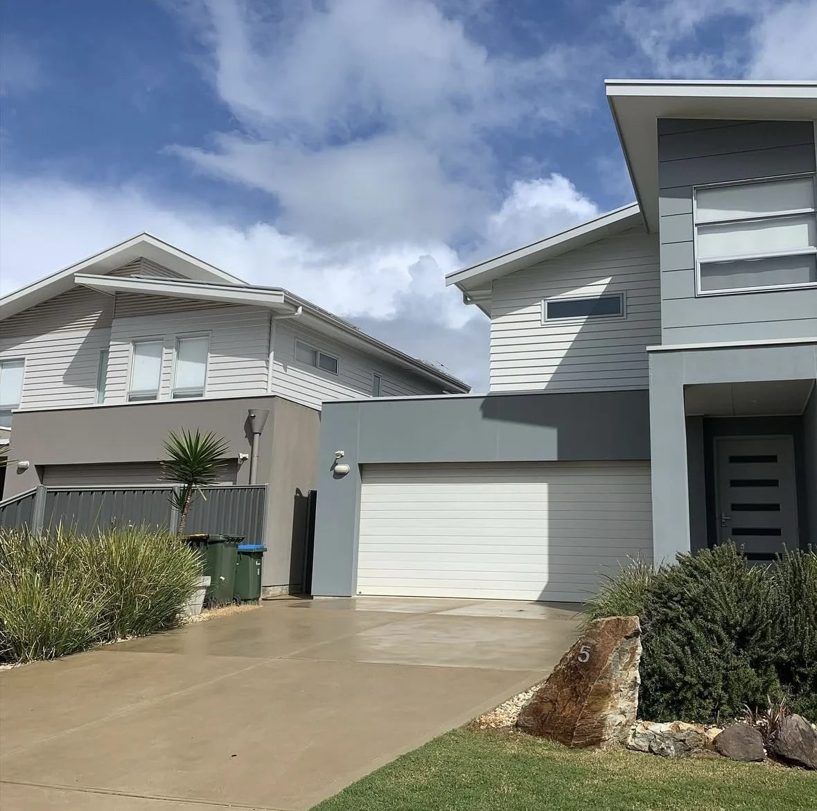 Modern two-story houses with gray and white exterior, driveway, garage, and landscaping under a partly cloudy sky.
