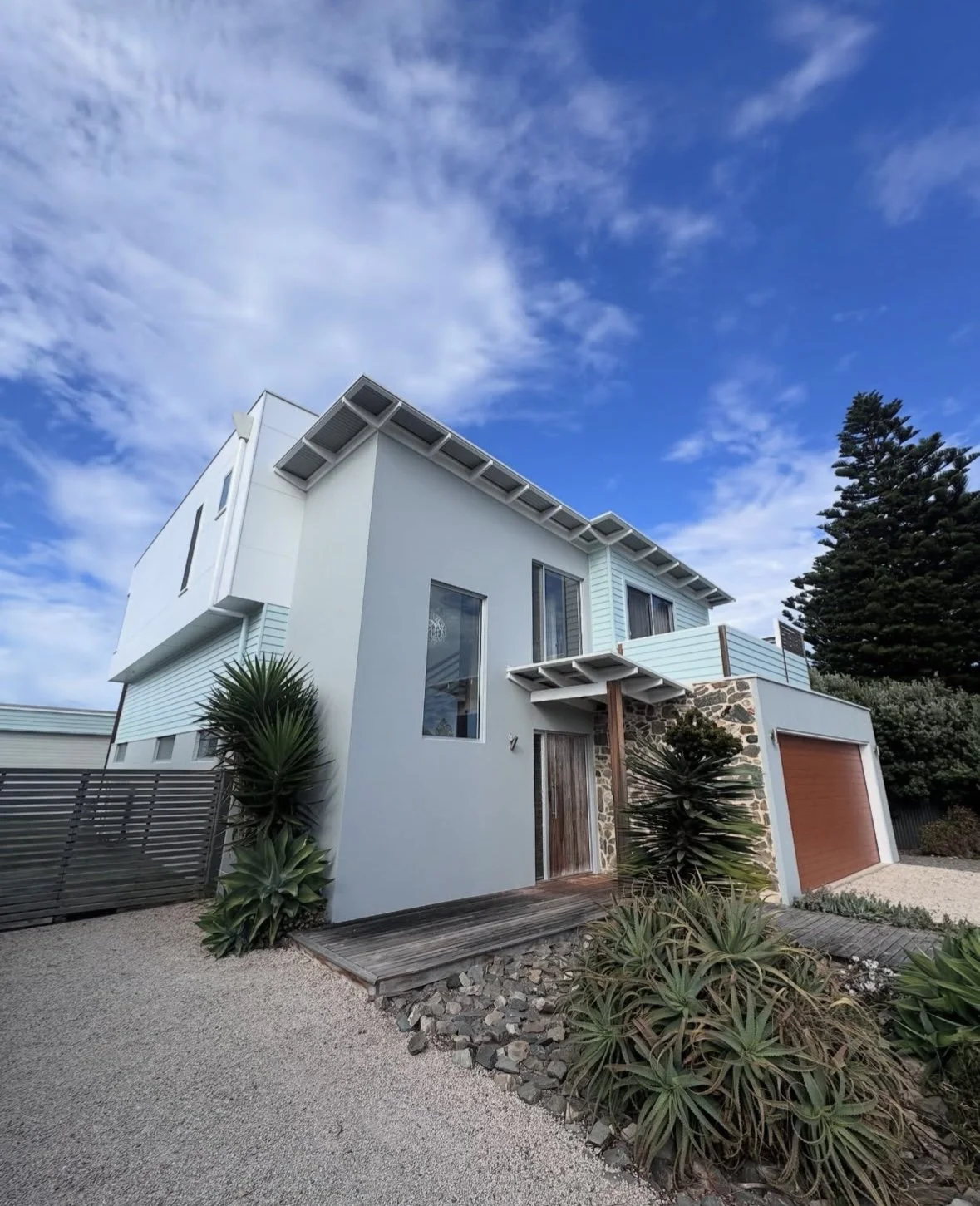 expertly painted Modern multi-story house with white walls, a wooden door, stone accents, and a garage door, surrounded by desert plants and gravel landscaping under a partly cloudy sky.