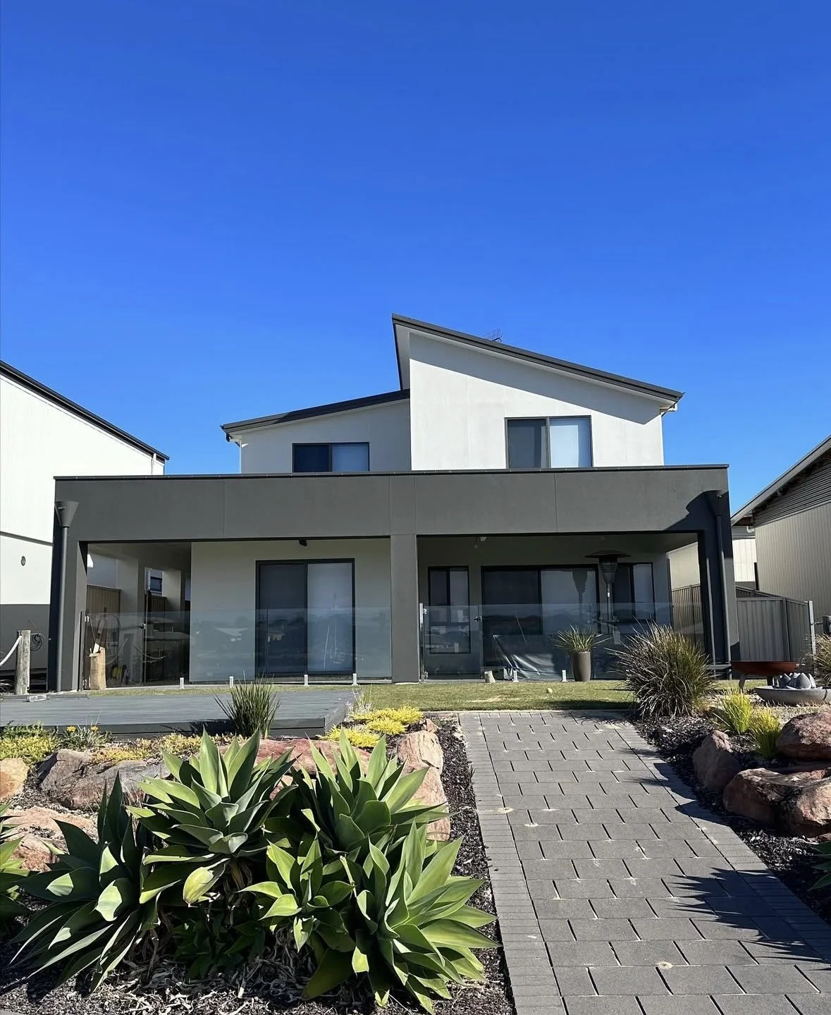 Modern two-story house with a white and dark gray exterior, slanted roof, large windows, glass sliding doors, and a landscaped front yard with plants, rocks, and a paved walkway.
