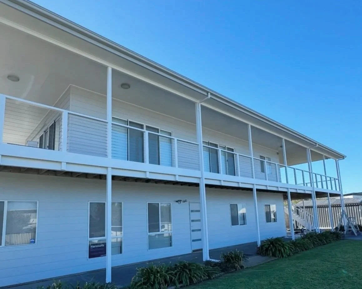freshly painted white multi-level residential building with a large balcony, glass sliding doors, small windows, and a grassy area with plants in front.