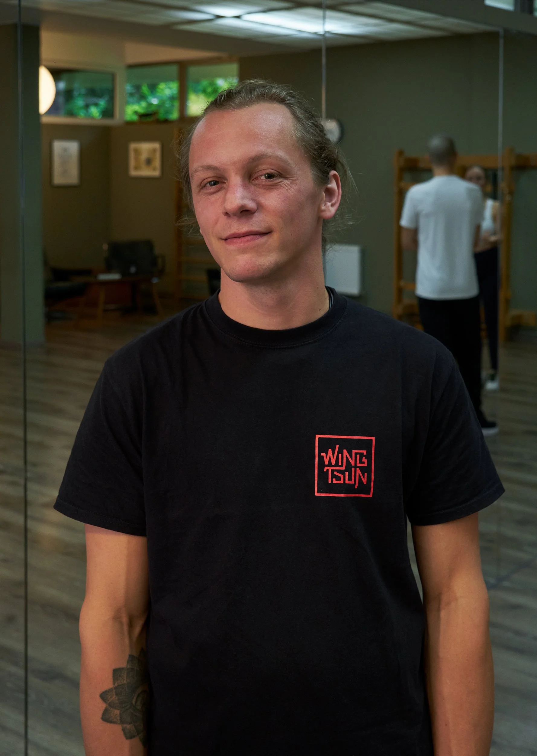 Ein junger Mann mit langen Haaren und einem Tattoo am linken Arm steht in einem Raum mit Spiegeln, trägt ein schwarzes T-Shirt mit rotem Schriftzug. Im Hintergrund sind andere Menschen und Yoga- oder Fitness-Equipment zu sehen.