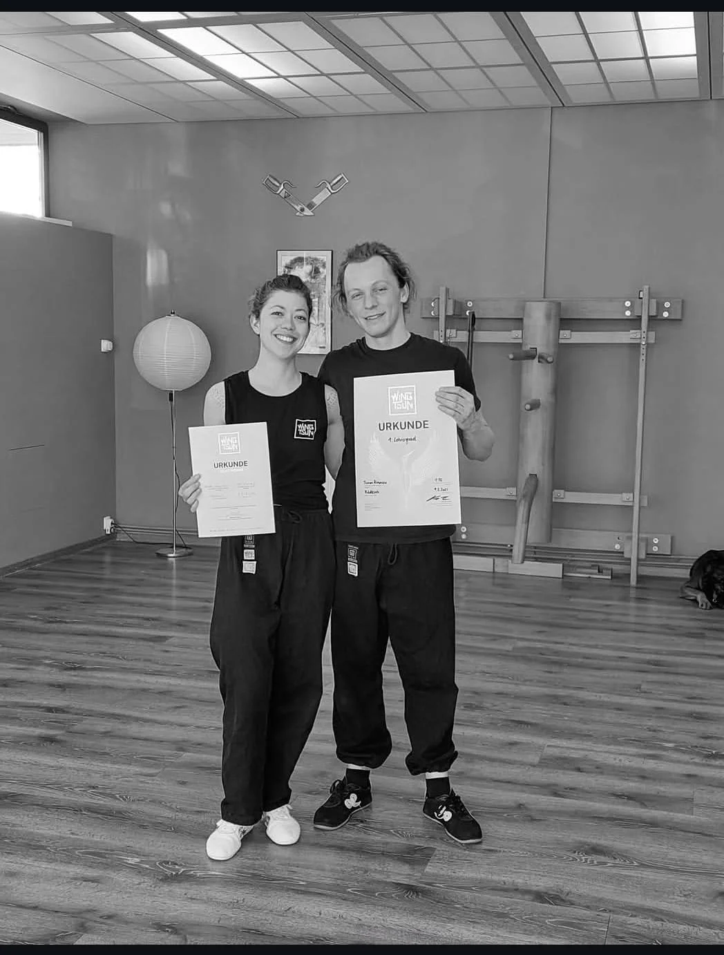 Zwei Personen, die in einem Fitnessstudio oder Kampfsportraum stehen, lächeln und halten Urkunden. Es sind Matten und Trainingsgeräte im Hintergrund sichtbar, eine Person trägt ein Wandei-Logo auf seinem T-Shirt.