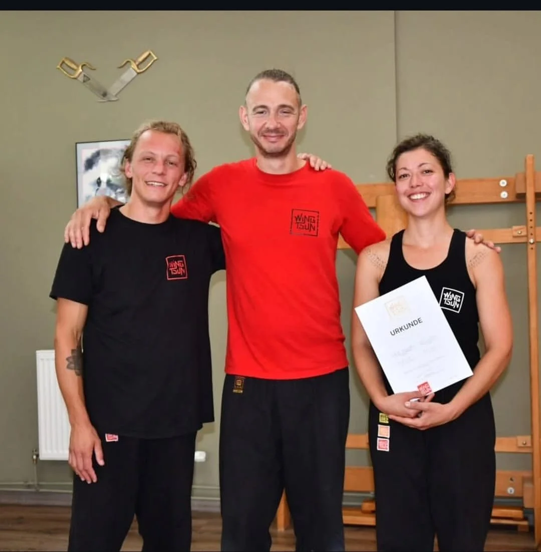 Drei Menschen, die T-Shirts mit einem Logo tragen, stehen zusammen in einem Raum und lächeln. Die Frau auf der rechten Seite hält eine Urkunde. Es scheint eine feierliche oder erfolgreiche Situation zu sein.