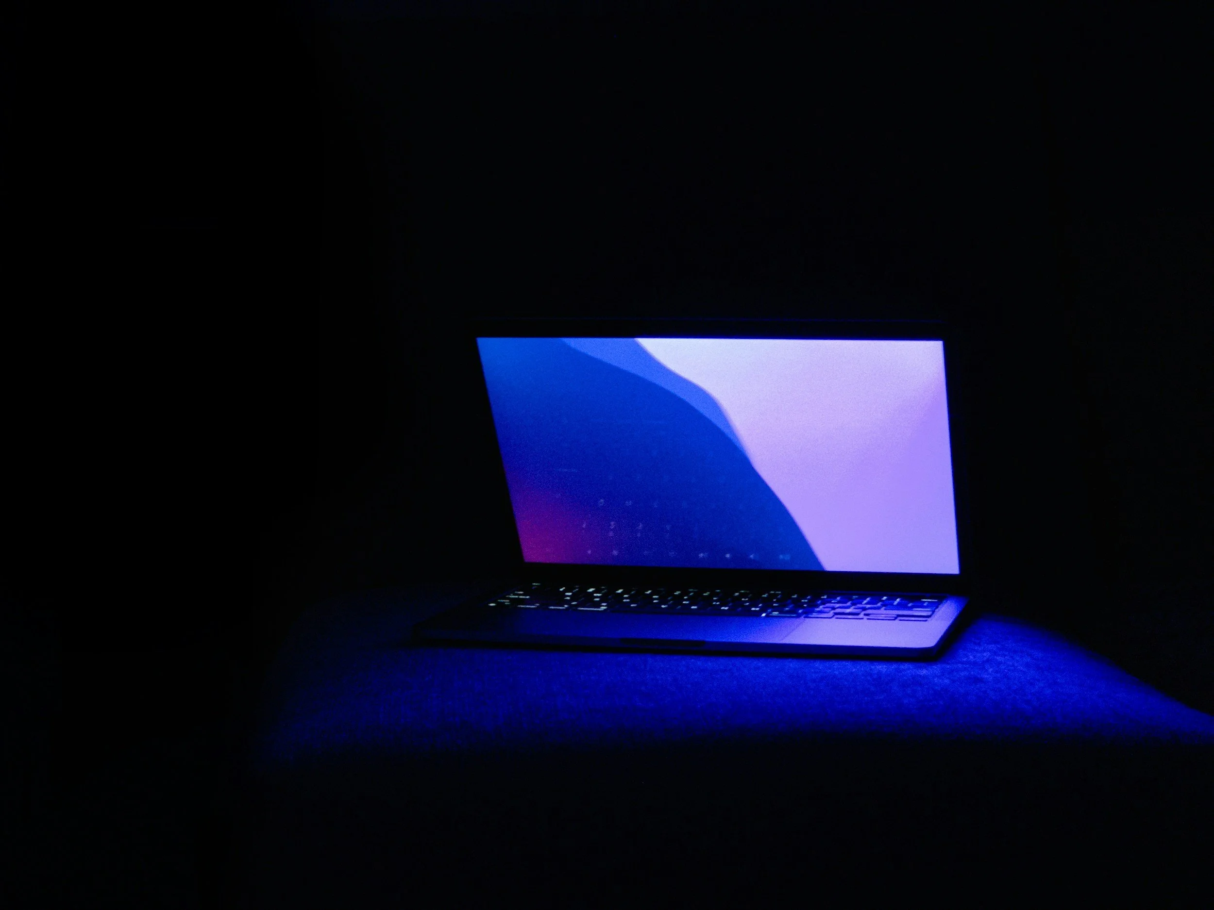 A laptop computer with a blue and purple gradient screen, illuminated in a dark room, sitting on a dark surface.