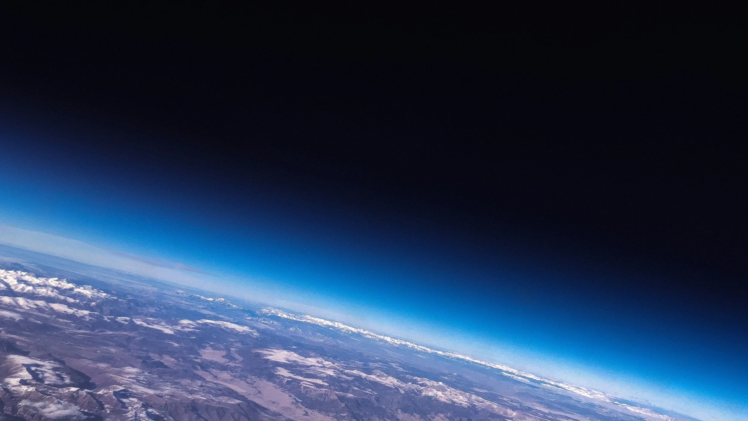 Aerial view of Earth's surface with snow-capped mountains and a dark upper atmosphere.