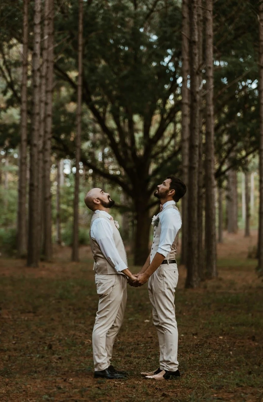 Two men in wedding attire holding hands and looking at each other in a forest with tall trees and a large central tree in the background.