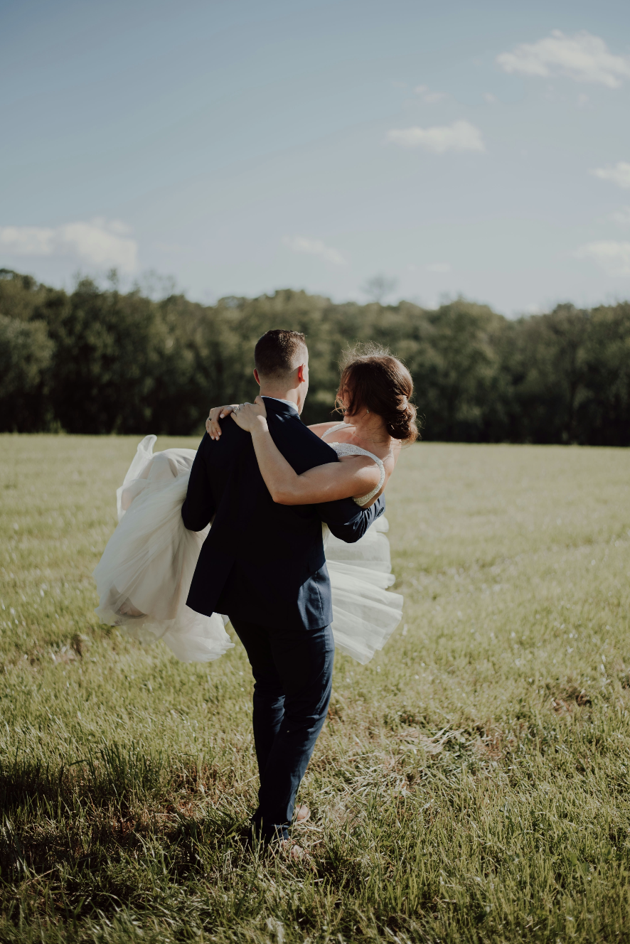 A groom in a dark suit carries a bride in a white wedding dress across a grassy field with trees in the background on a sunny day.