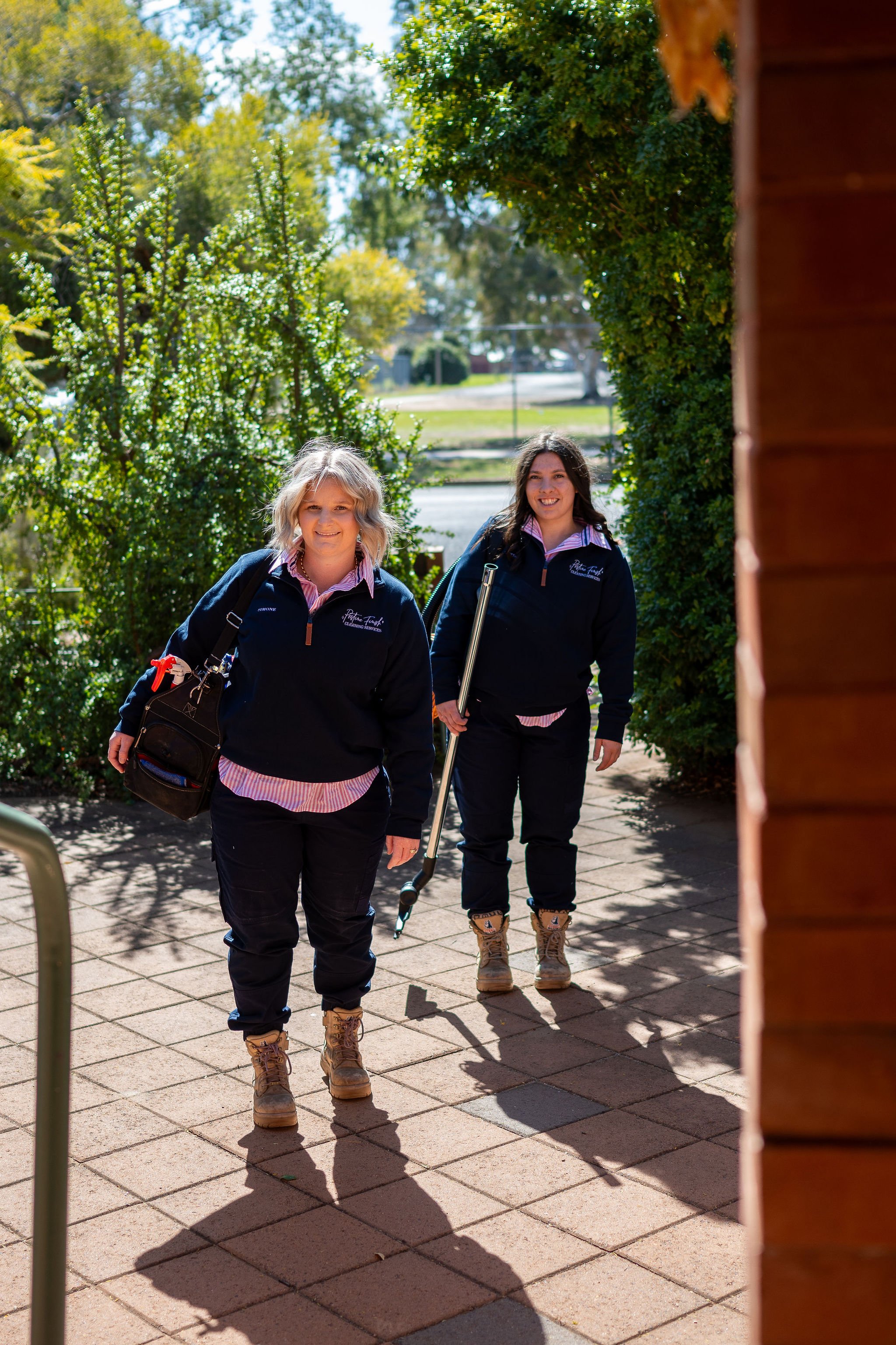 Pristine Finish Cleaning Staff wearing matching dark navy jackets with pink and white striped shirts underneath, walking outside on a paved path surrounded by greenery, smiling at the camera, with shadows cast on the ground.